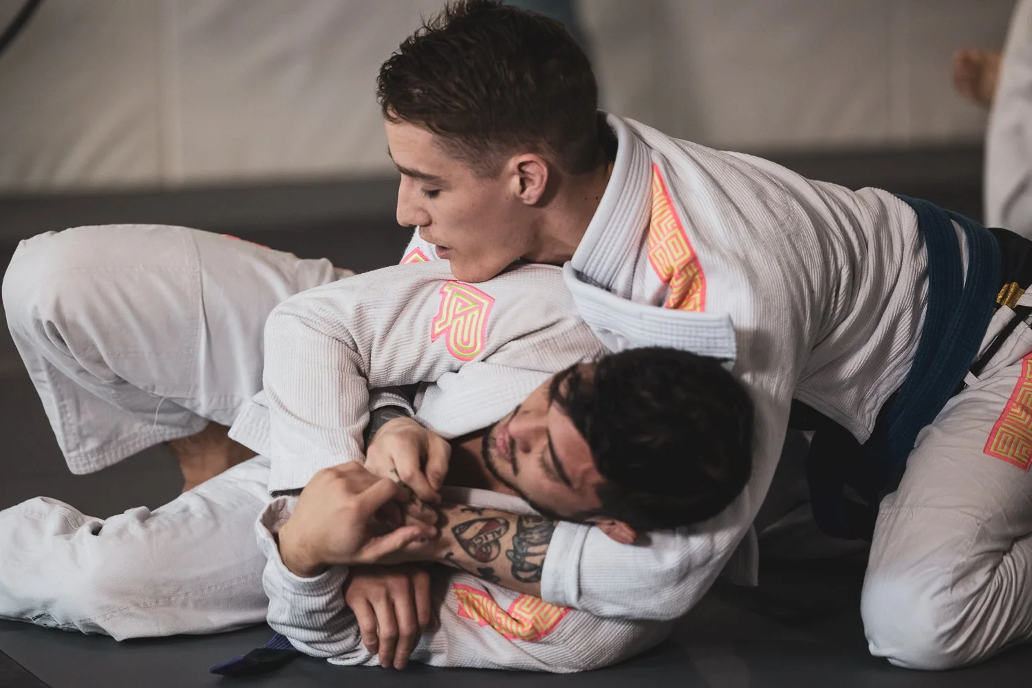 Two males practicing Brazilian Jiu-Jitsu, wearing gis, on a mat during training.