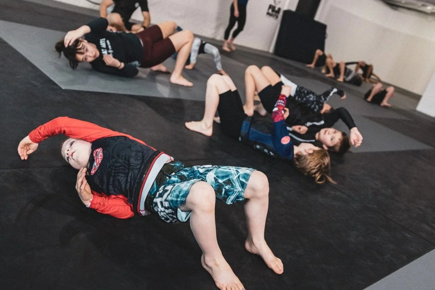 Children and adults practicing Brazilian Jiu-Jitsu on mats in a martial arts gym, performing various grappling positions.