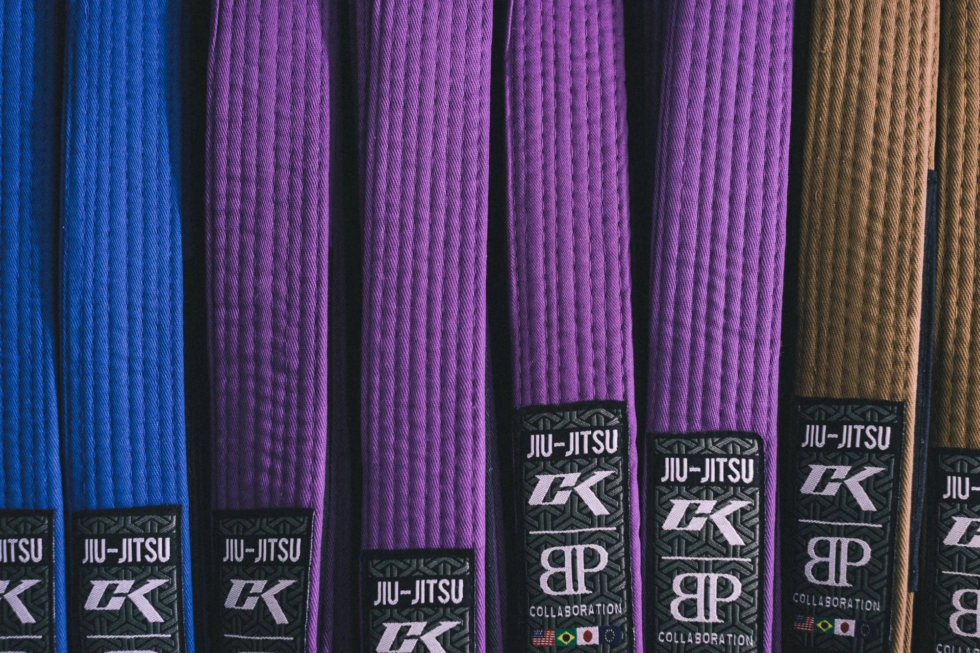 Colored martial arts belts in blue, purple, brown, and black with Jiu-Jitsu and collaboration logos at the bottom.