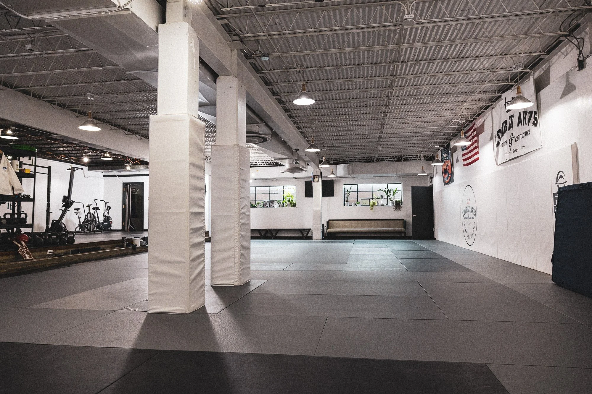 Interior of a martial arts gym with mats on the floor, white walls, and a large banner reading "Combat Arts" with American flag and gym equipment visible.