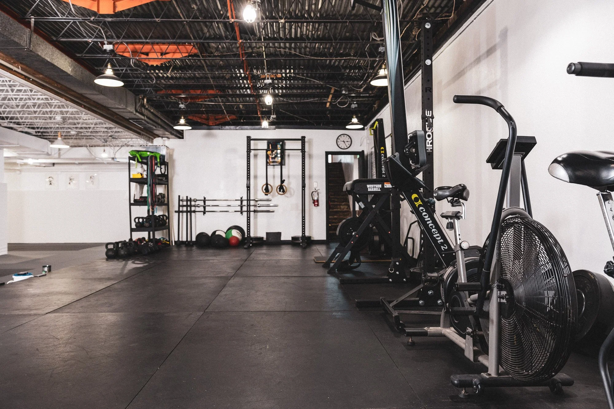 Empty gym with workout equipment including stationary bikes, kettlebells, medicine balls, battle ropes, and exercise machines, with a black ceiling, white walls, and a wall clock.