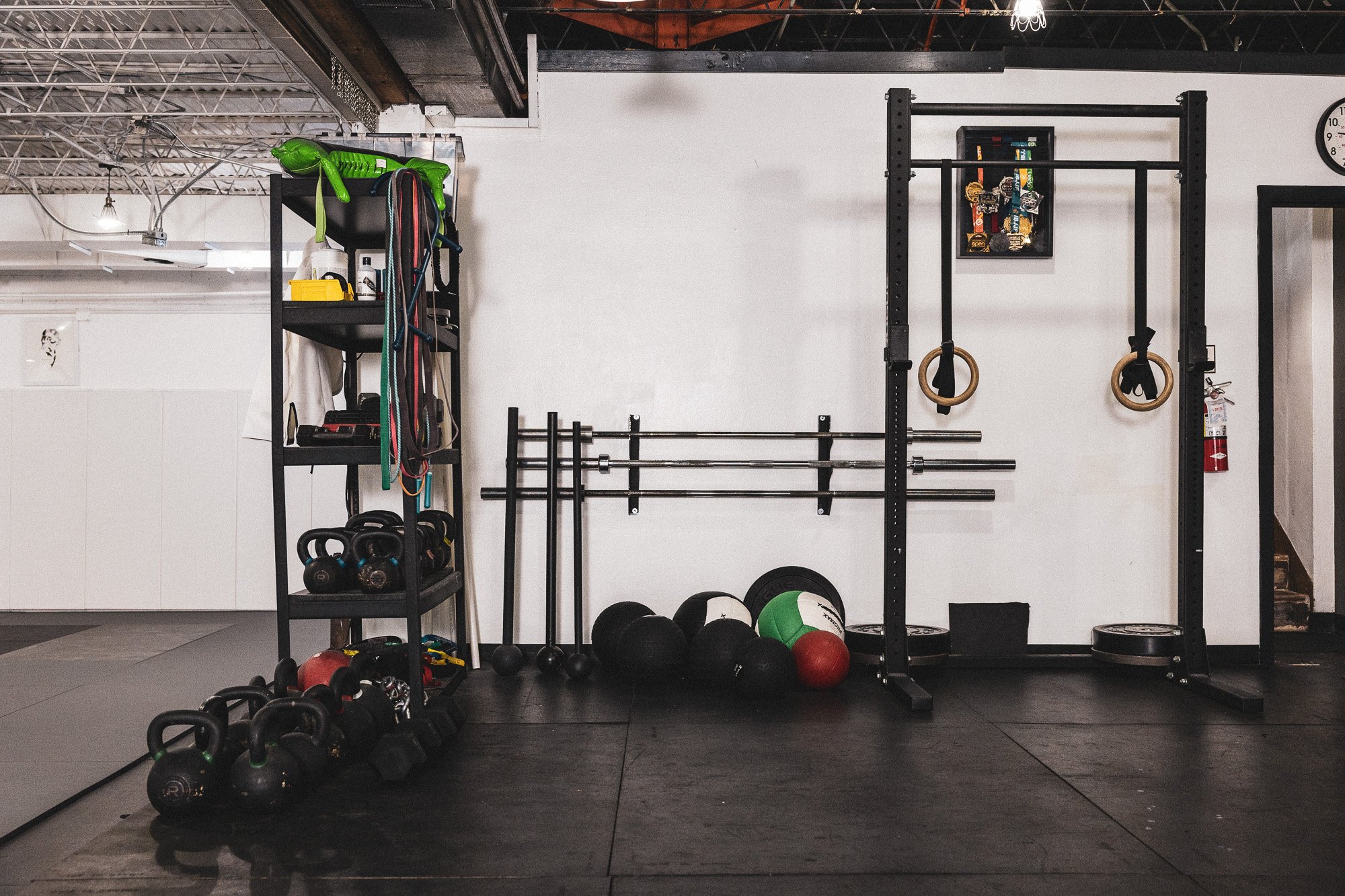 A gym corner with black metal shelving holding colorful resistance bands, bottles, and other small gym equipment. Below there's a row of kettlebells on the floor, along with medicine balls. On the wall, a black rack holds barbells and gymnastic rings