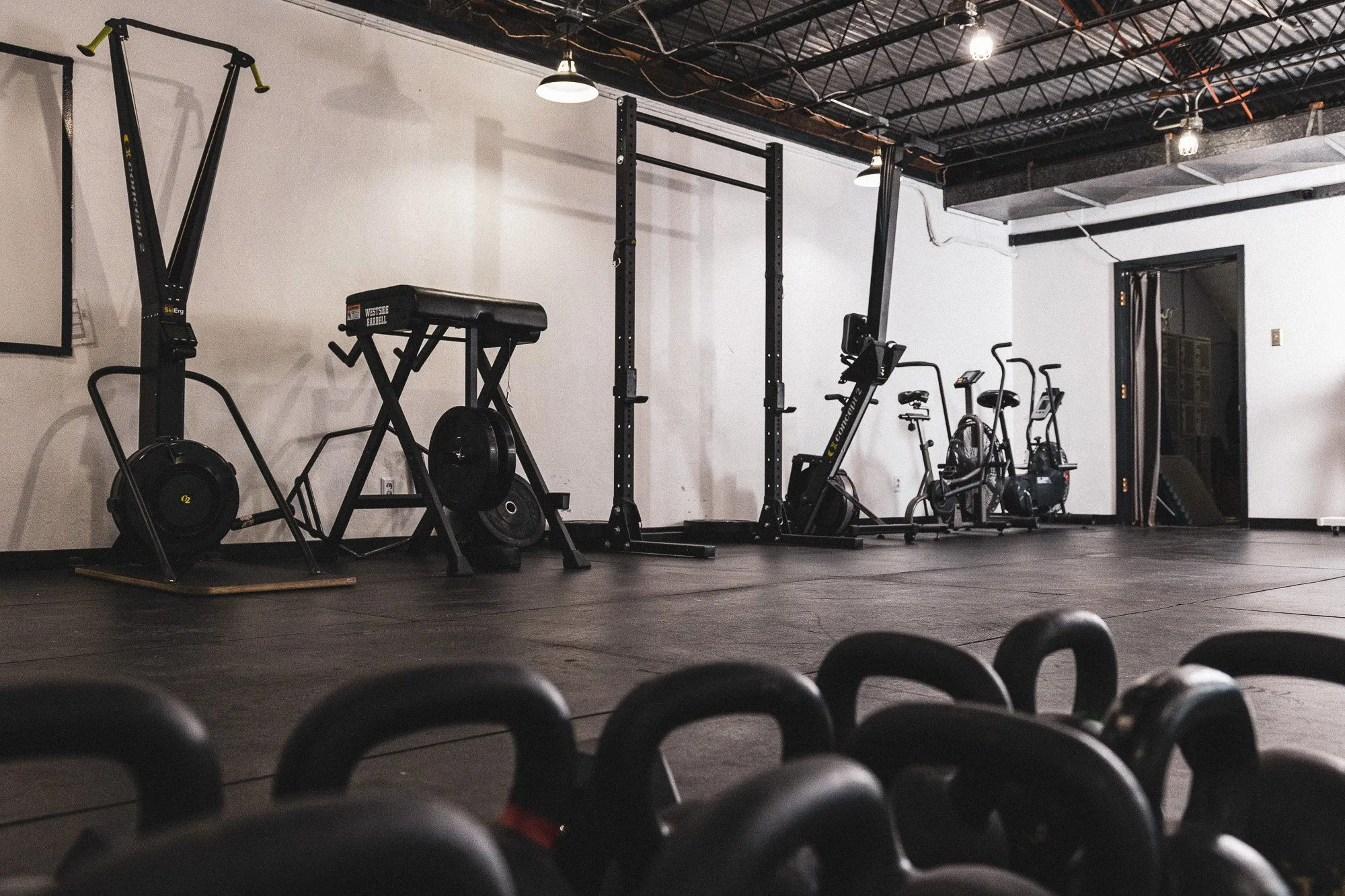 Interior of a gym with black exercise equipment, including stationary bikes, rowing machines, and a squat rack. The gym has black flooring, white walls, exposed ceiling pipes, and hanging industrial lights.