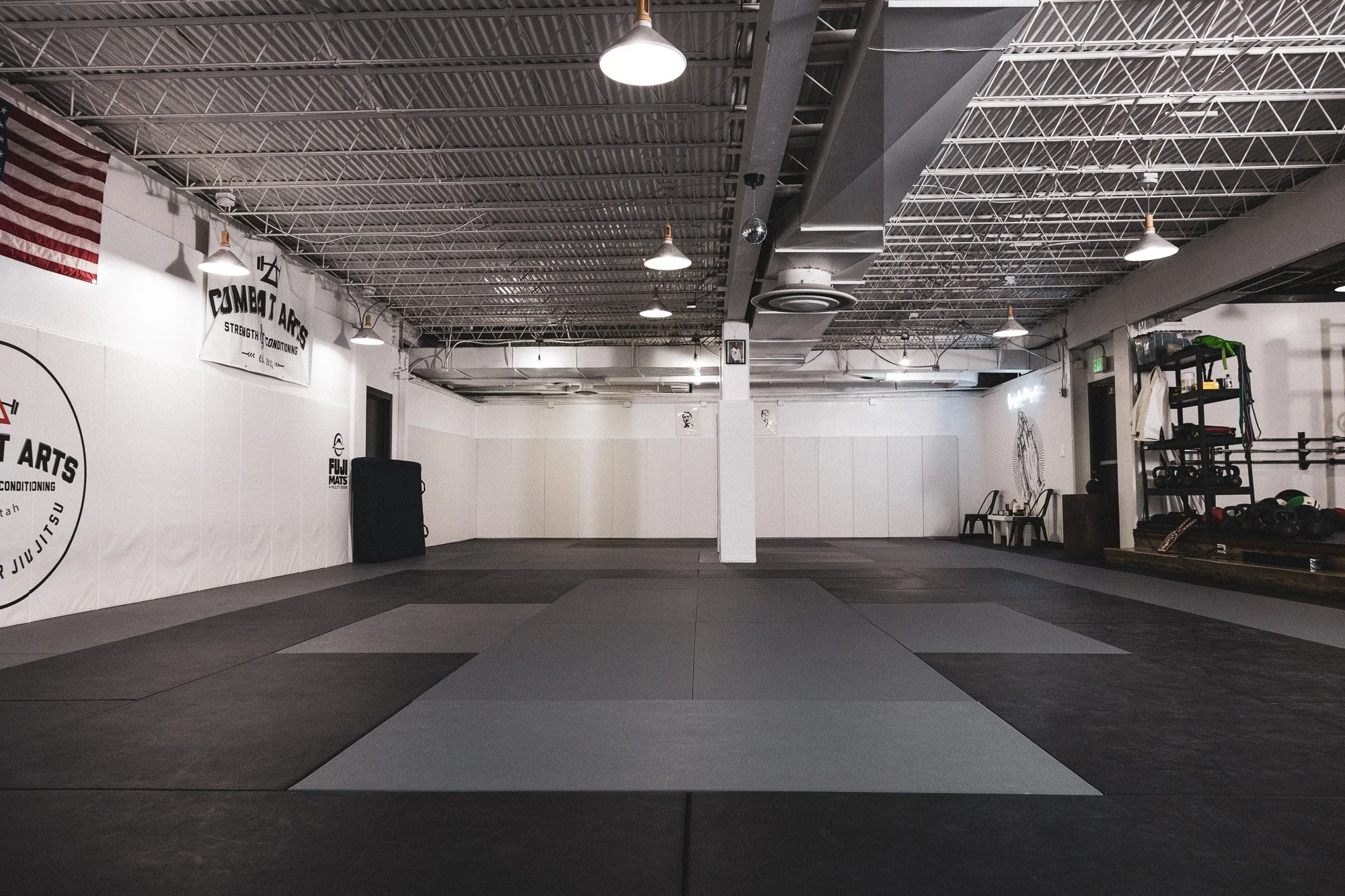 An interior view of a martial arts training facility with a large open mat area, white walls, and fitness equipment on the right side. The ceiling features exposed metal beams and hanging lights.