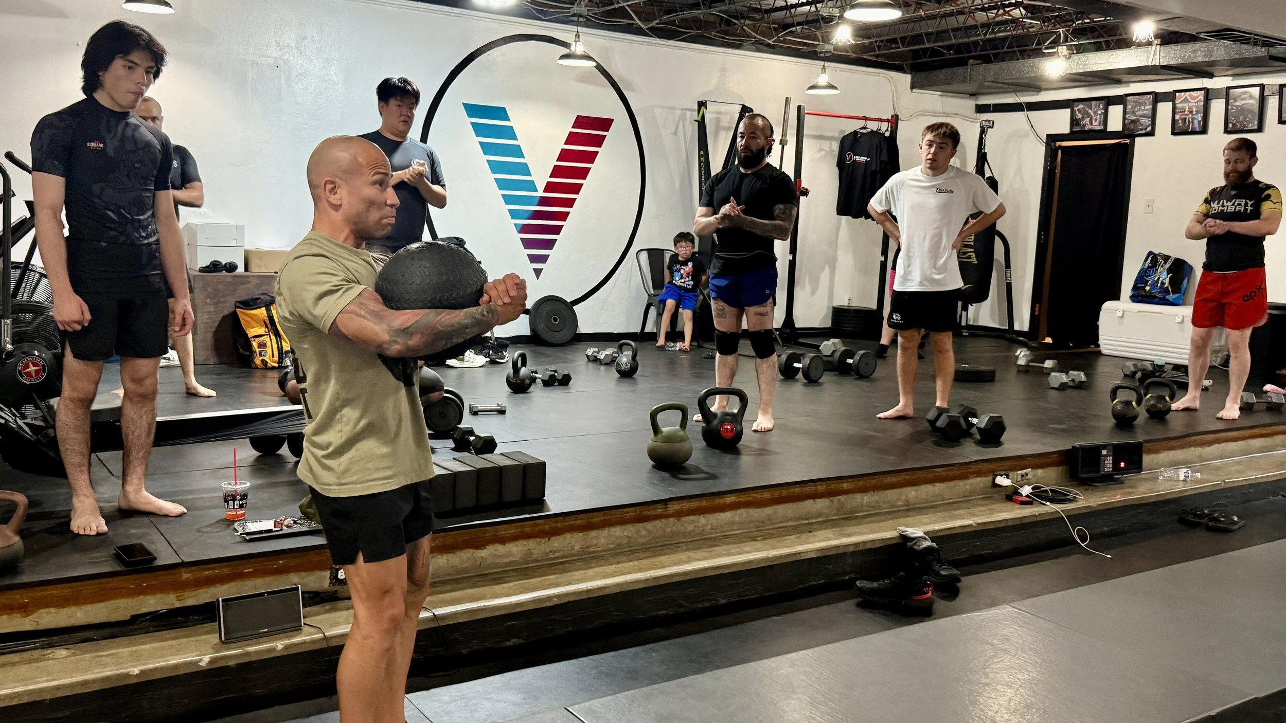Group of people in a Group Strength and conditioning fitness class, some standing and one holding a medicine ball, with a large logo on the wall behind them, gym equipment scattered on the floor, and framed photos on the wall.