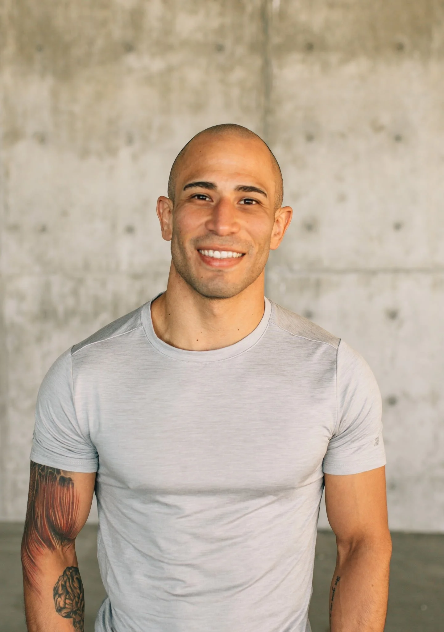 A smiling man with a shaved head wearing a light gray t-shirt, standing in front of a concrete wall.