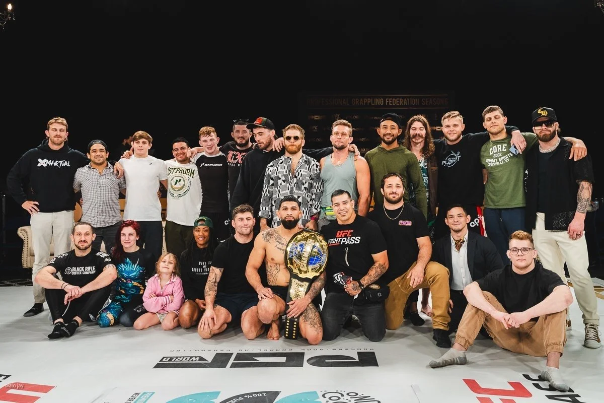 A large group of people, including a mixed group of men, women, and children, posing together in an MMA fighting ring. The central figure is a man holding a championship belt, sitting on the mat.