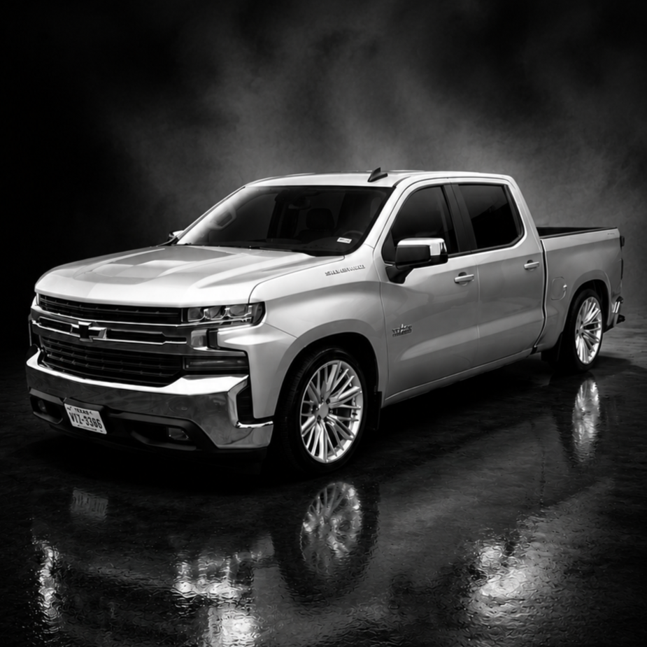 A silver Chevrolet Silverado pickup truck parked in a studio with reflections on the wet floor and a dark smoky background.