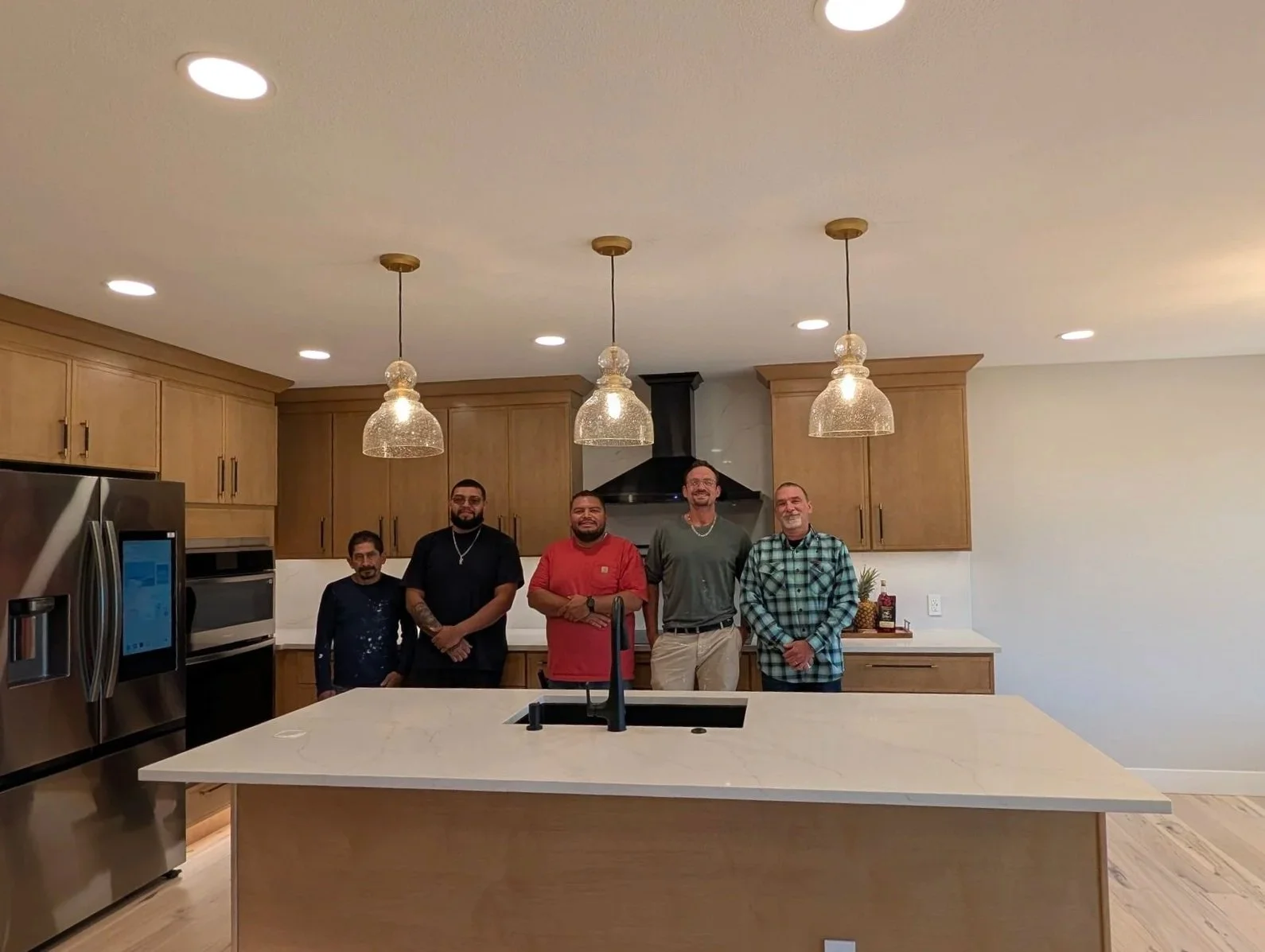 Five men standing in a modern kitchen with wooden cabinets, stainless steel appliances, hanging pendant lights, and a large white island counter.