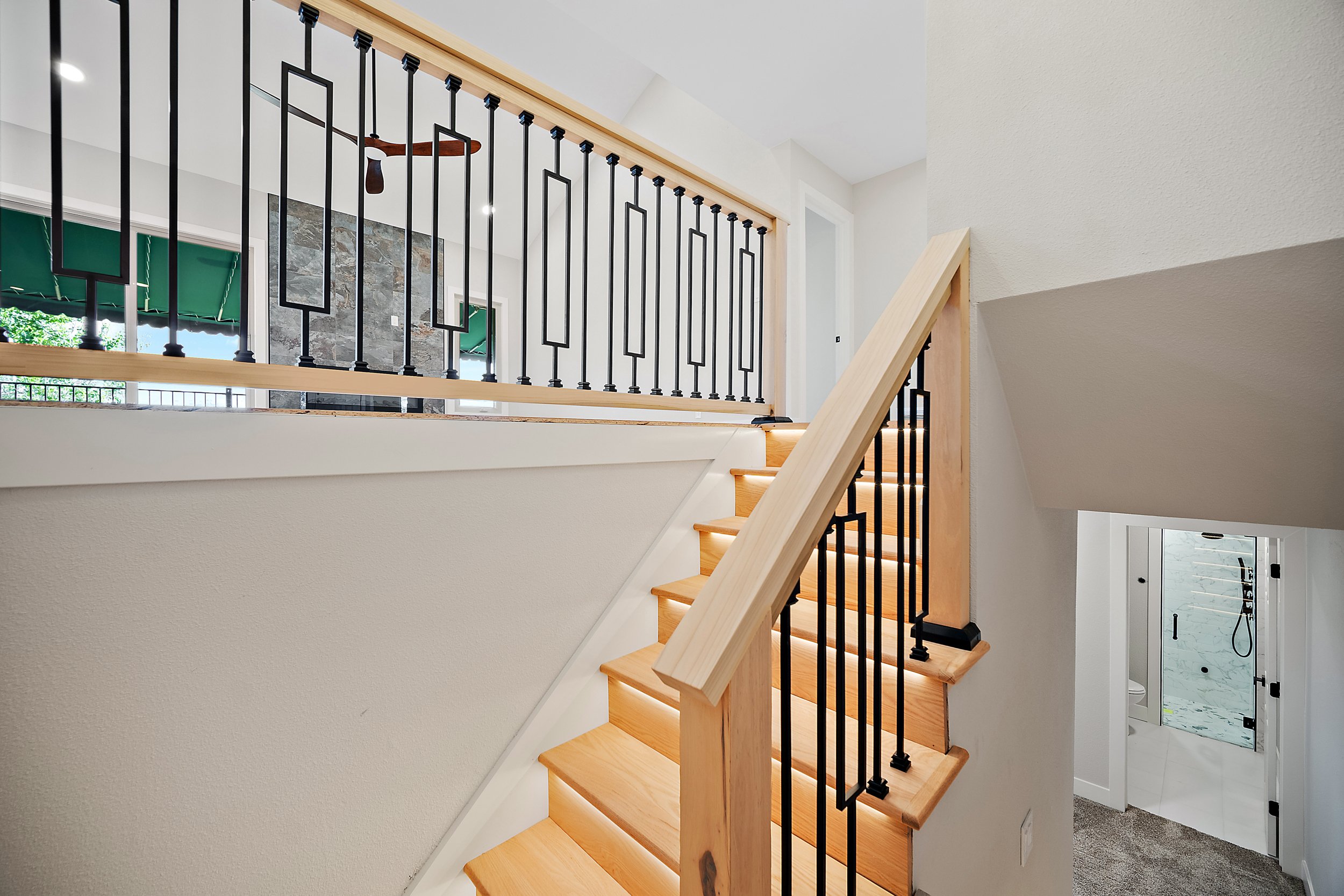 Interior view of a staircase with wooden steps, a light wooden handrail, and black metal balusters, leading to a second floor with visible windows and a stone accent wall in the background.