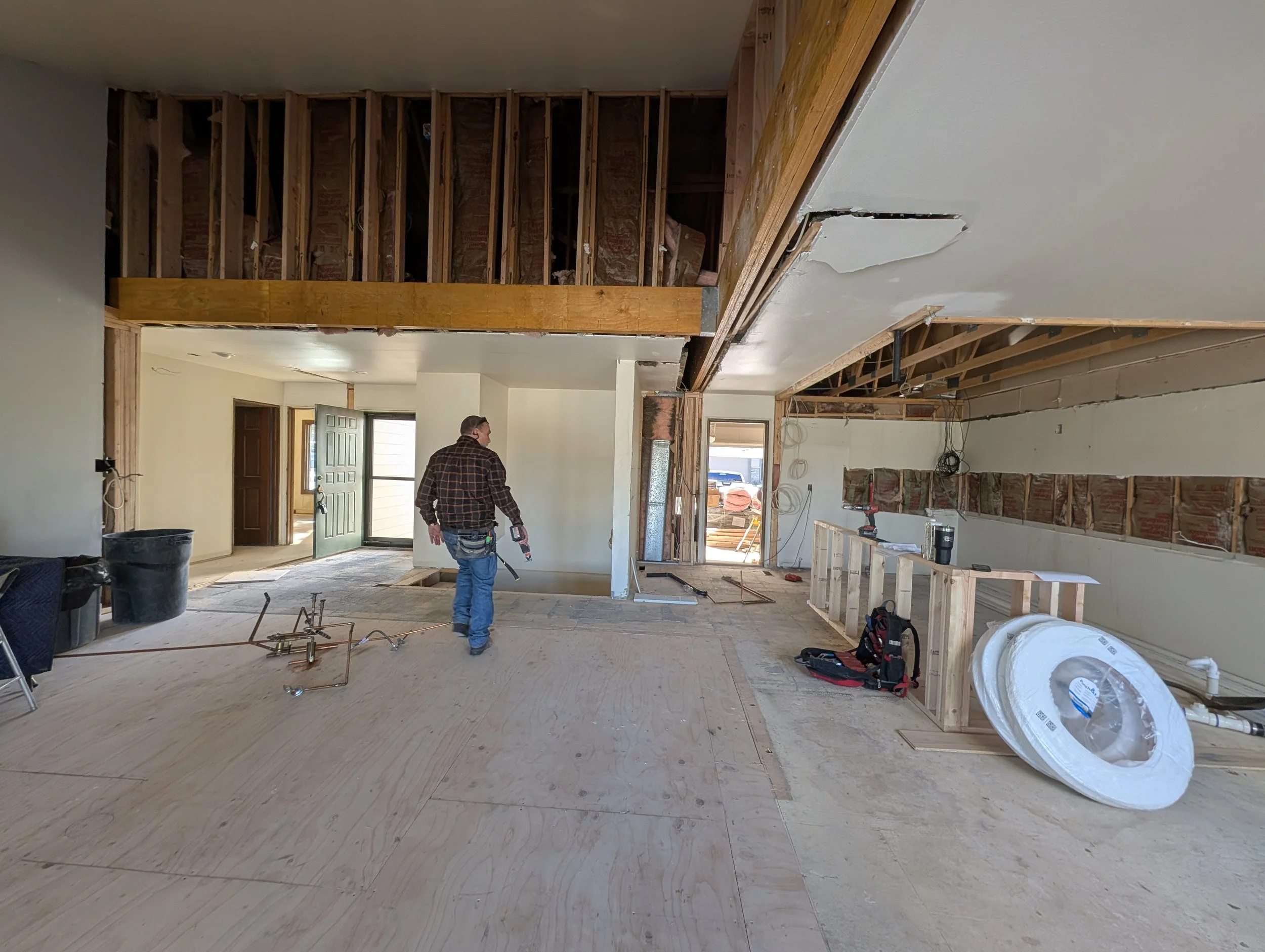 Interior of a house under renovation with exposed wall studs, a worker holding a tool, construction materials, and wiring visible.