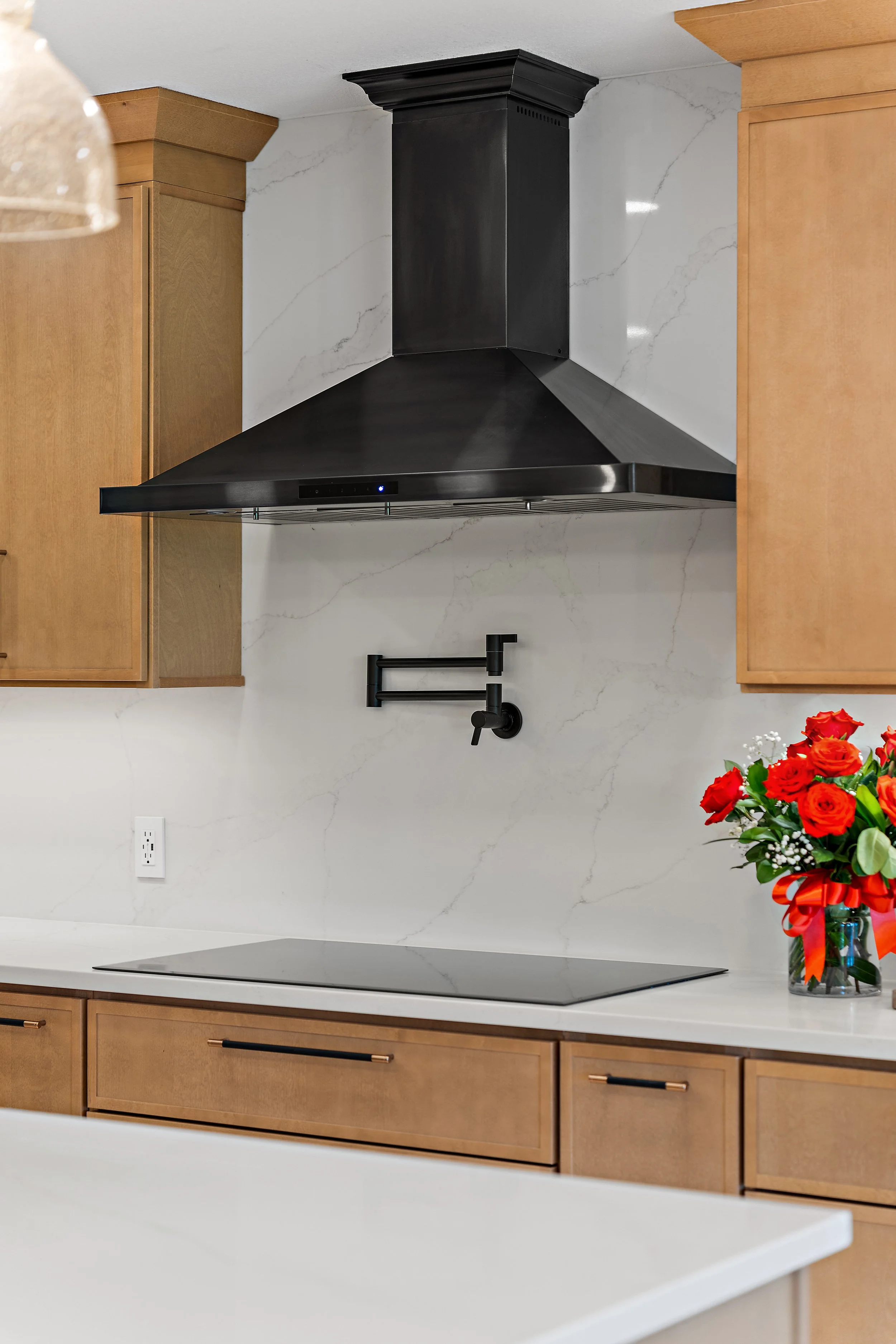 A modern kitchen with a black range hood, light wood cabinets, a white marble backsplash, a black faucet, white countertop, and a bouquet of red and white flowers in a glass vase with a red ribbon.