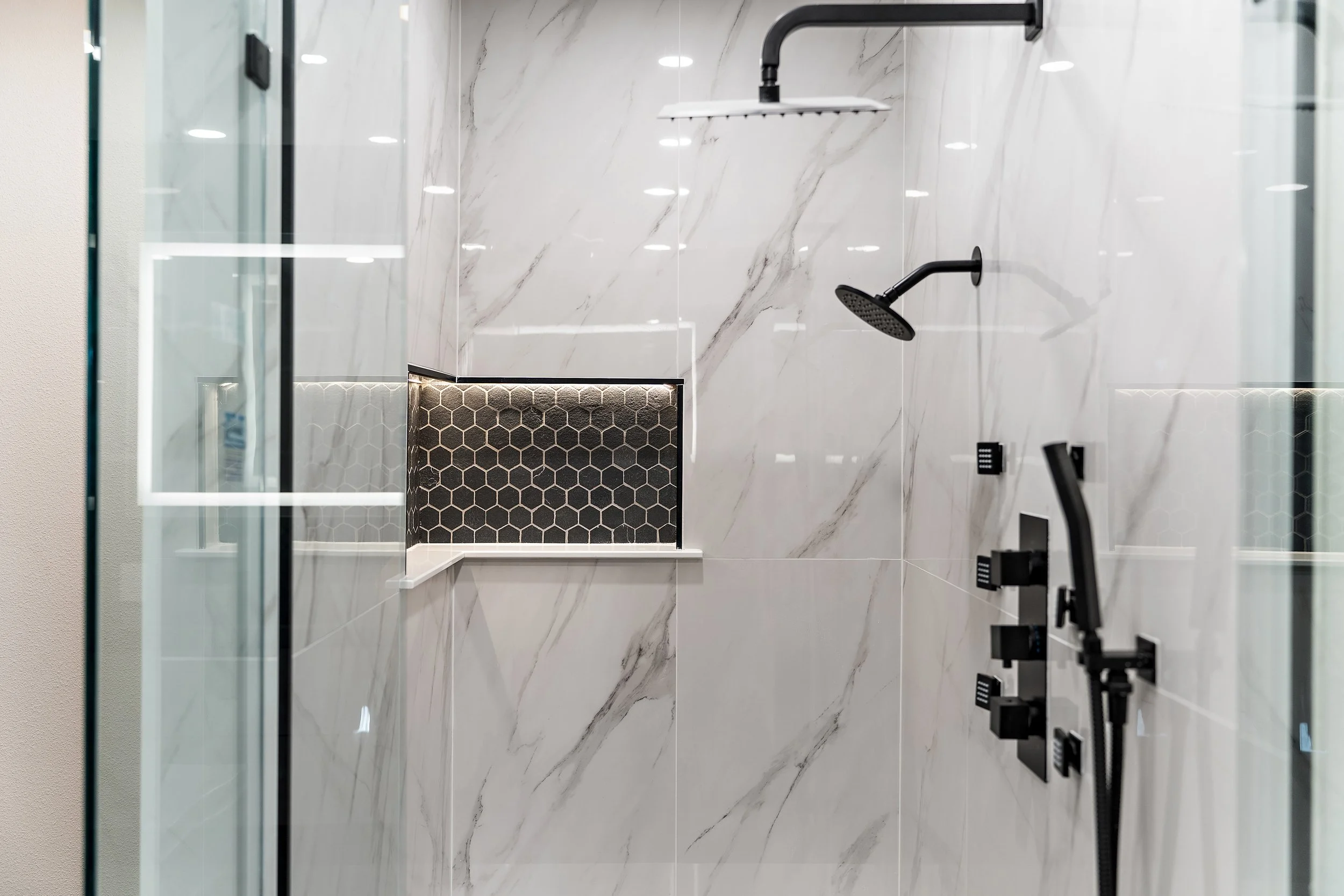 Modern shower area with white marbled walls, black showerhead, handheld shower, and black faucet fixtures, viewed through a glass partition.