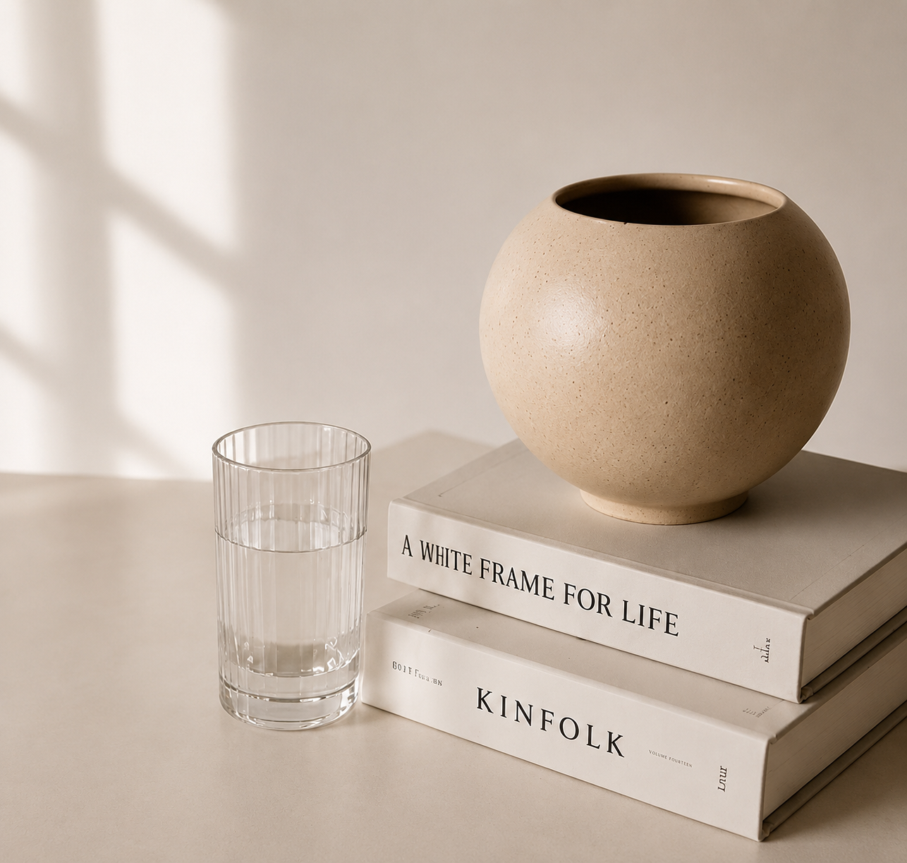 A beige round ceramic vase placed on top of two stacked white books titled "A WHITE FRAME FOR LIFE" and "KINFOLK." Next to the books, there is a clear glass of water on a light-colored surface. Shadows from a window create a pattern on the wall in the background.