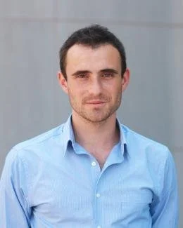 Portrait of a young man with short dark hair, wearing a light blue dress shirt, standing outdoors with a neutral background.