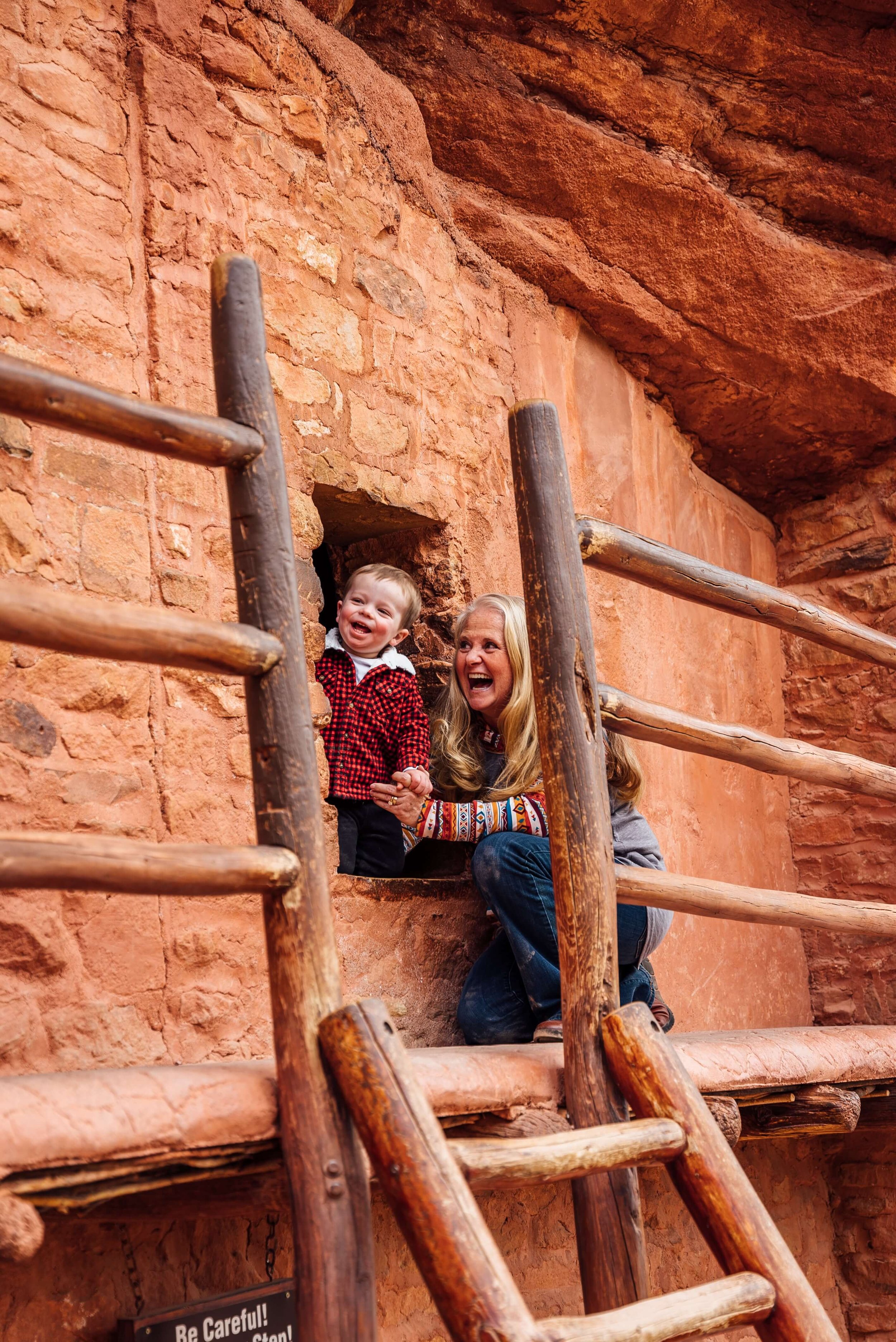 A woman and a young boy are peeking out of a small window in a red rock wall at the Manitou Cliff Dwellings in Manitou Springs, Colorado.