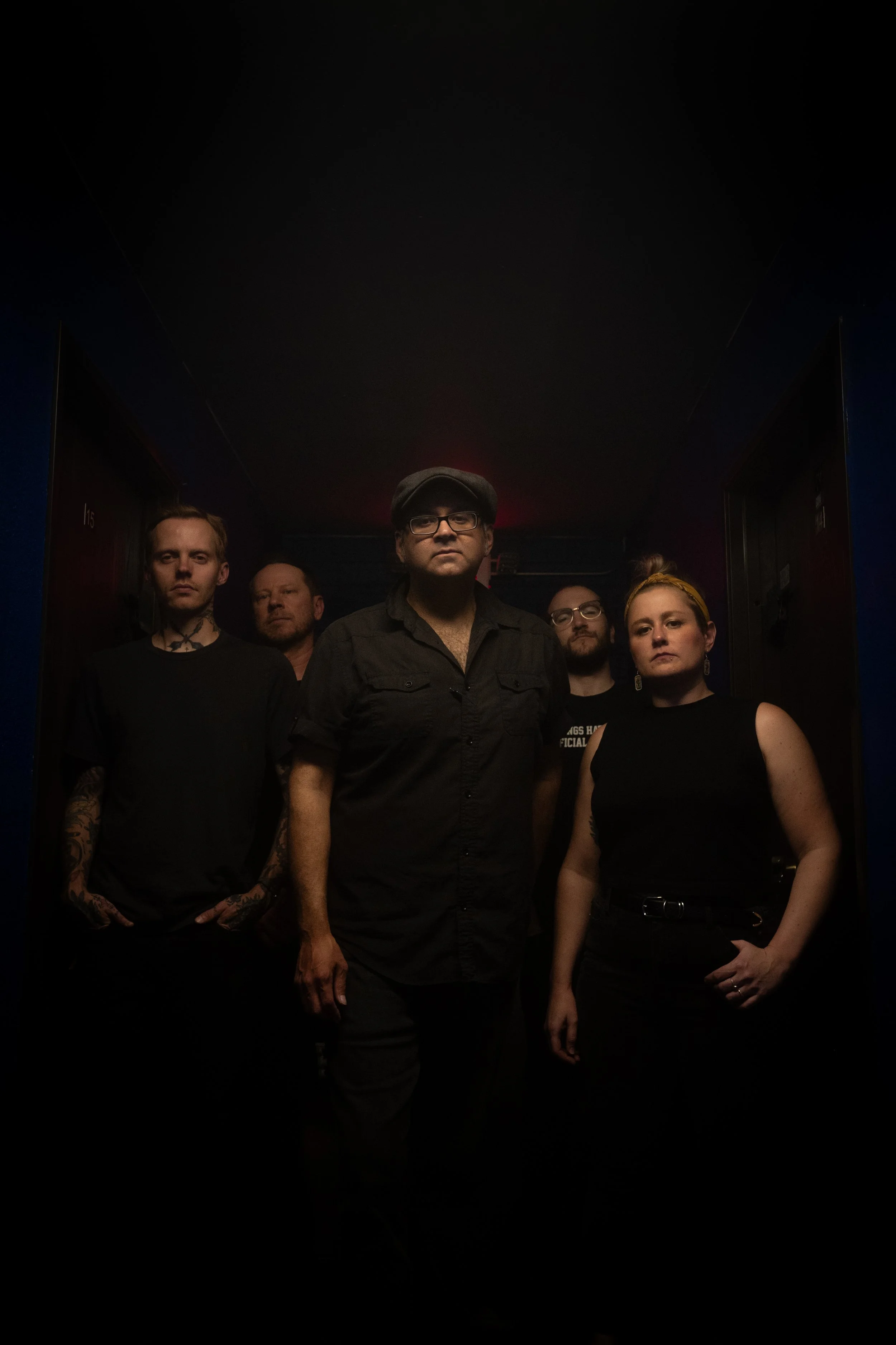 A group of five people standing in a dark room with minimal lighting, facing the camera with serious expressions.