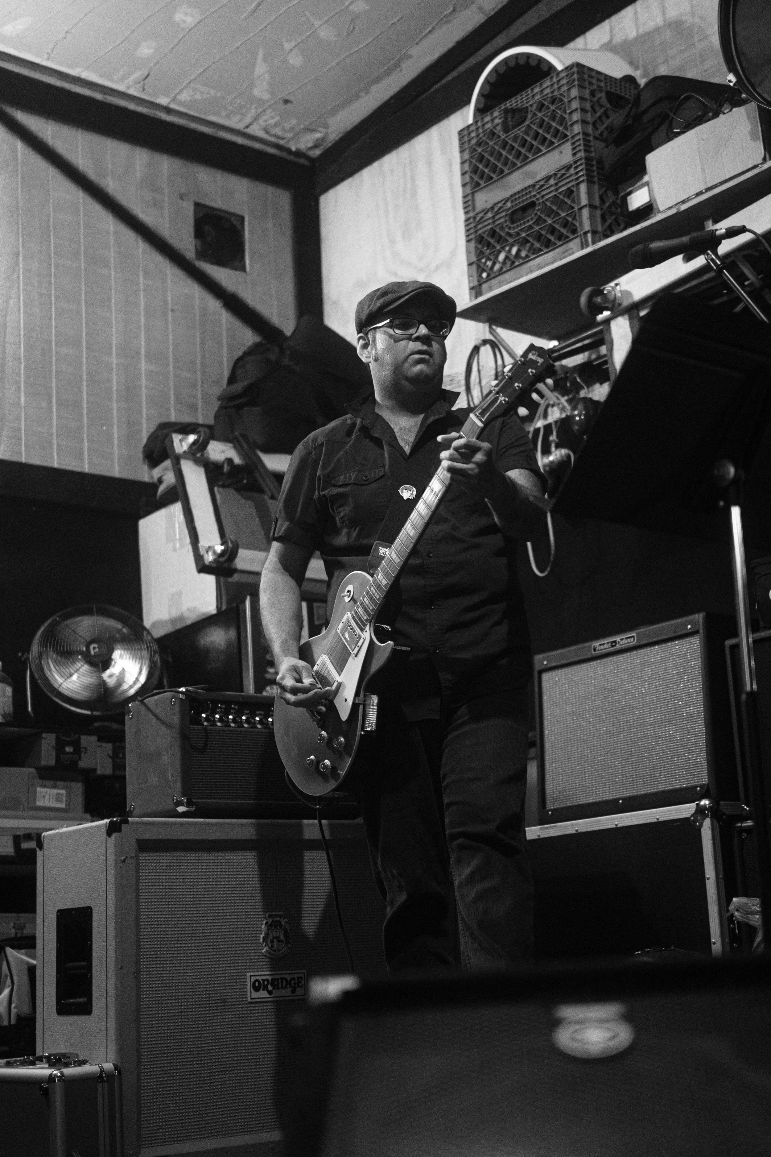A man playing an electric guitar in a rehearsal space or music studio, surrounded by amplifiers and equipment, in black and white.