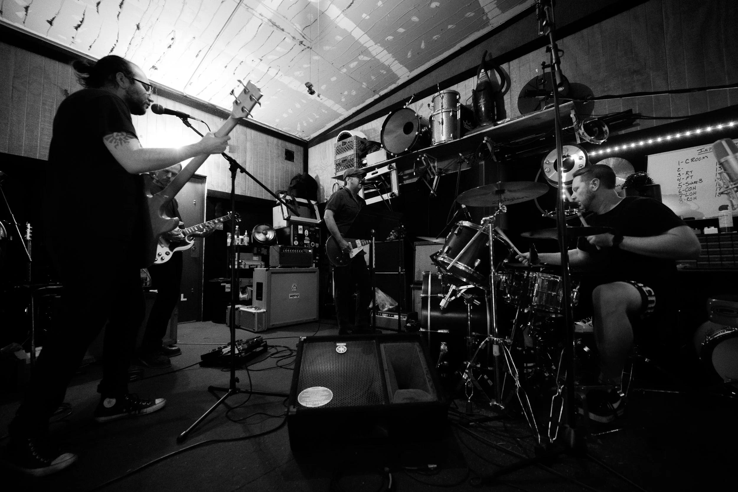 A black-and-white photo of a band rehearsing in a room with musical instruments, including guitars, drums, and a keyboard. Four band members are visible, with one playing guitar, another singing into a microphone, and another playing drums. The room 