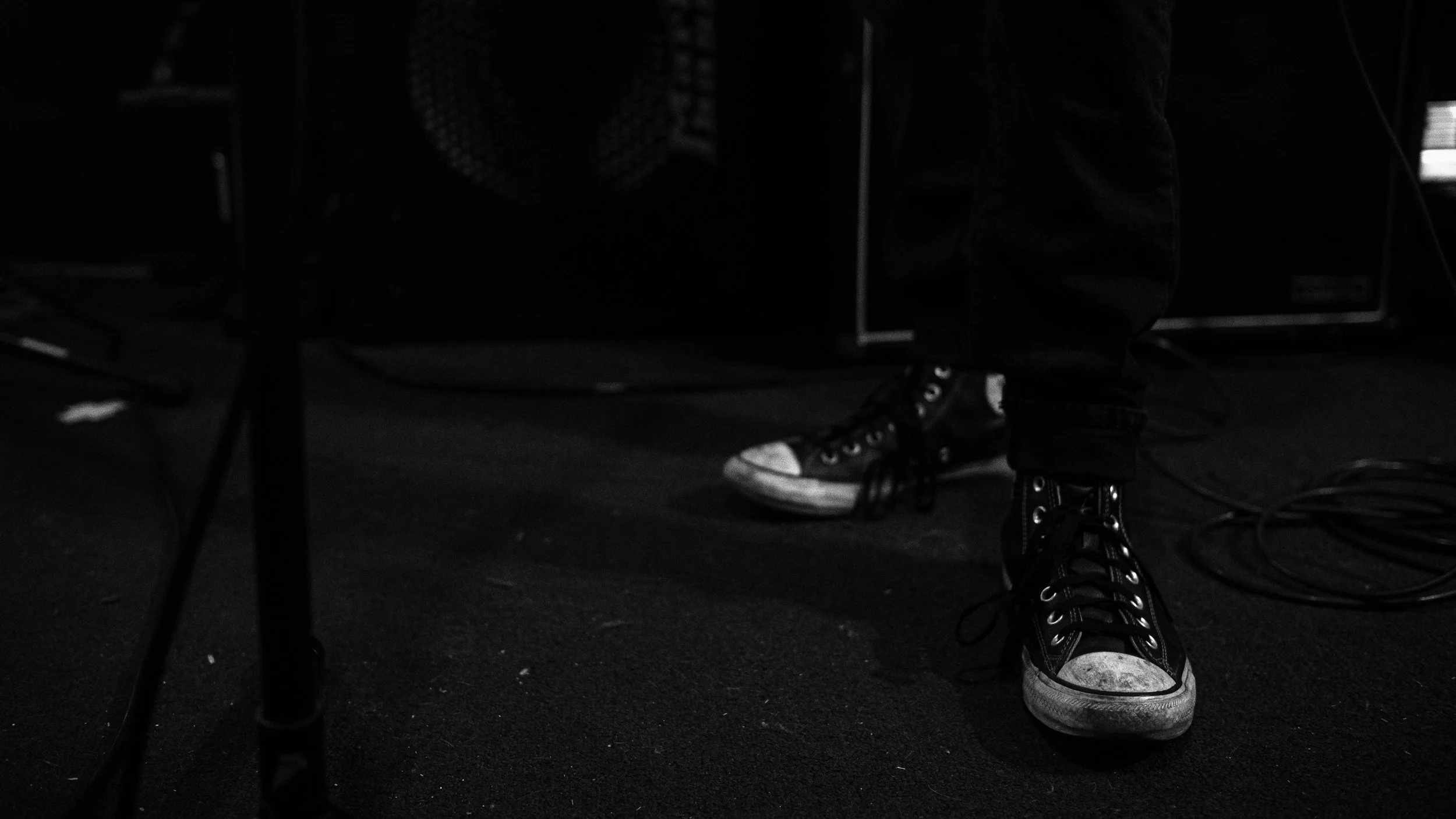 Close-up of a person's legs in black jeans and worn high-top sneakers, standing. Only the lower half of the person is visible, with a black and white sneakers, surrounded by cables and electronic equipment.