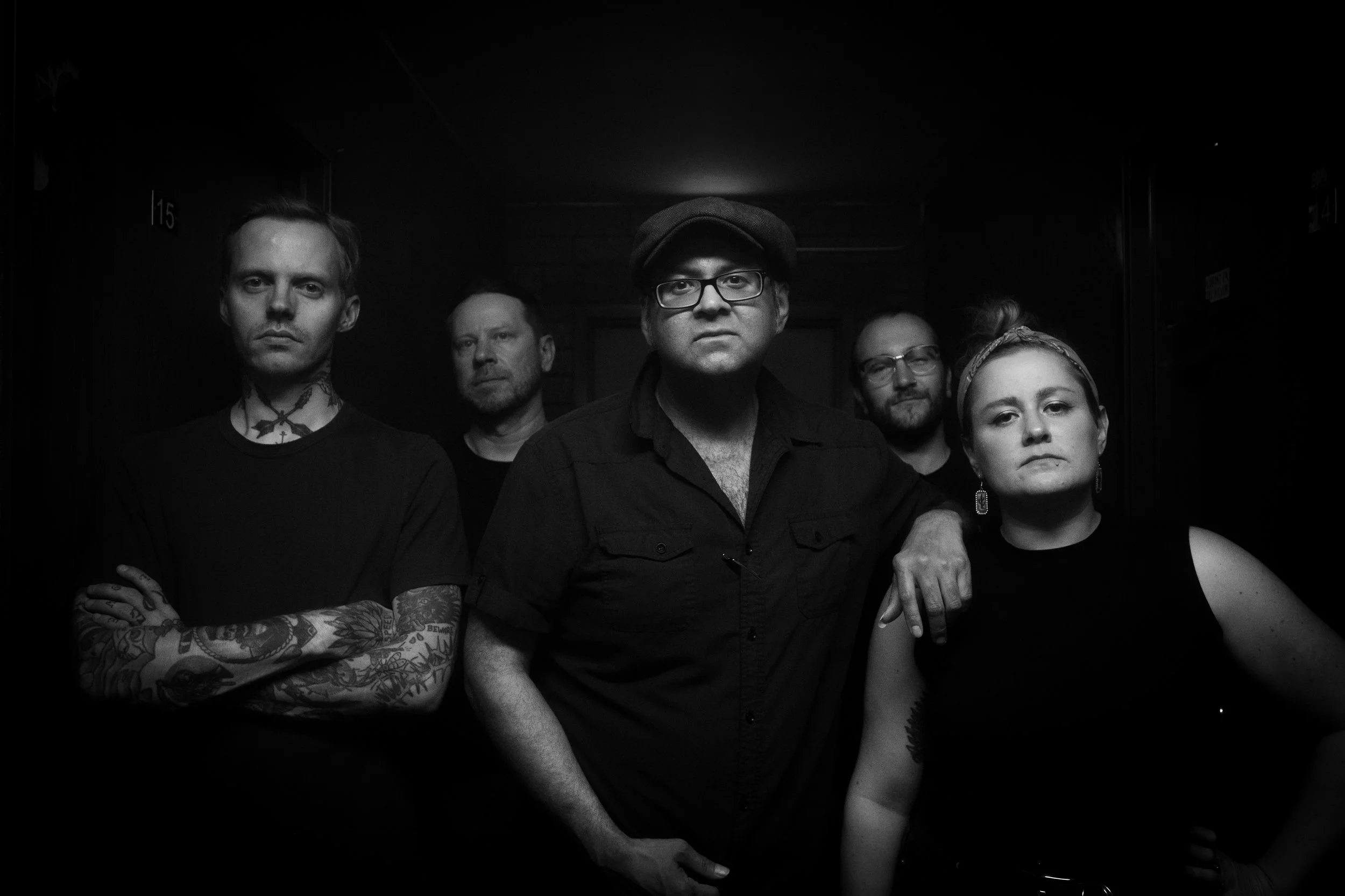 Group of five people standing together in a dimly lit space, with serious expressions. 
