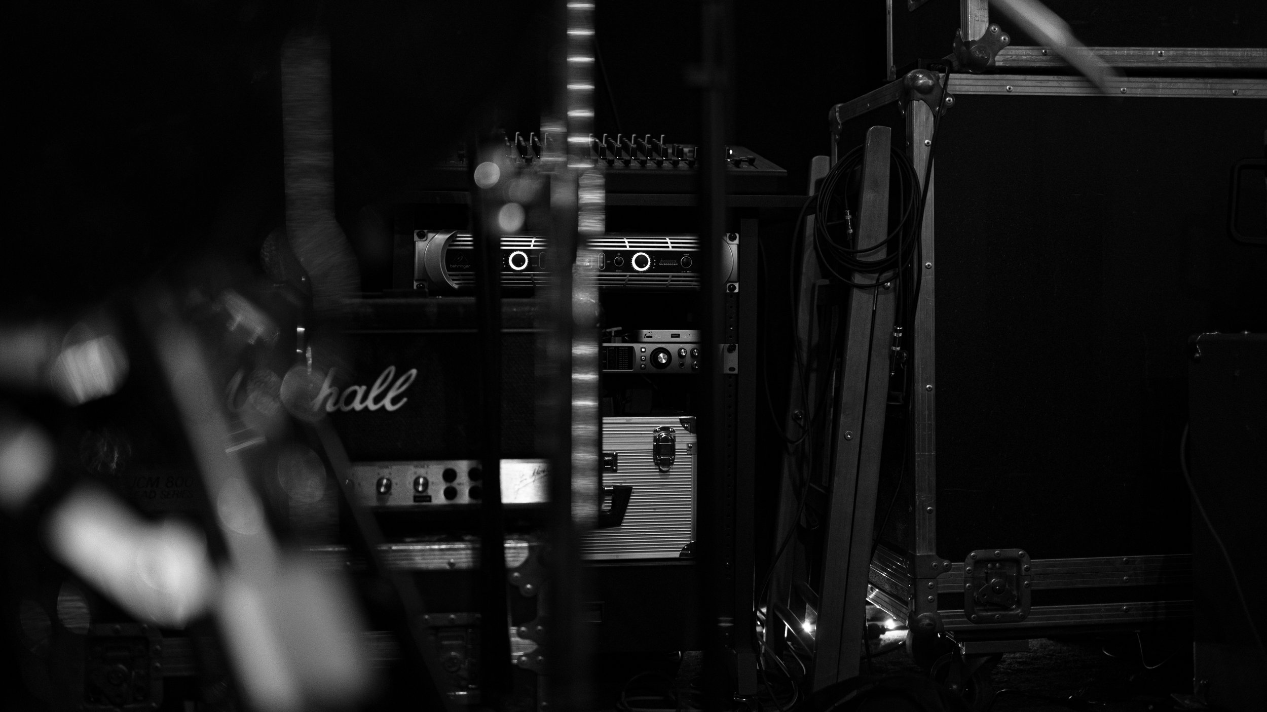 Black and white photo of audio equipment, including amplifiers, cables, and sound gear, set up in a dark space.