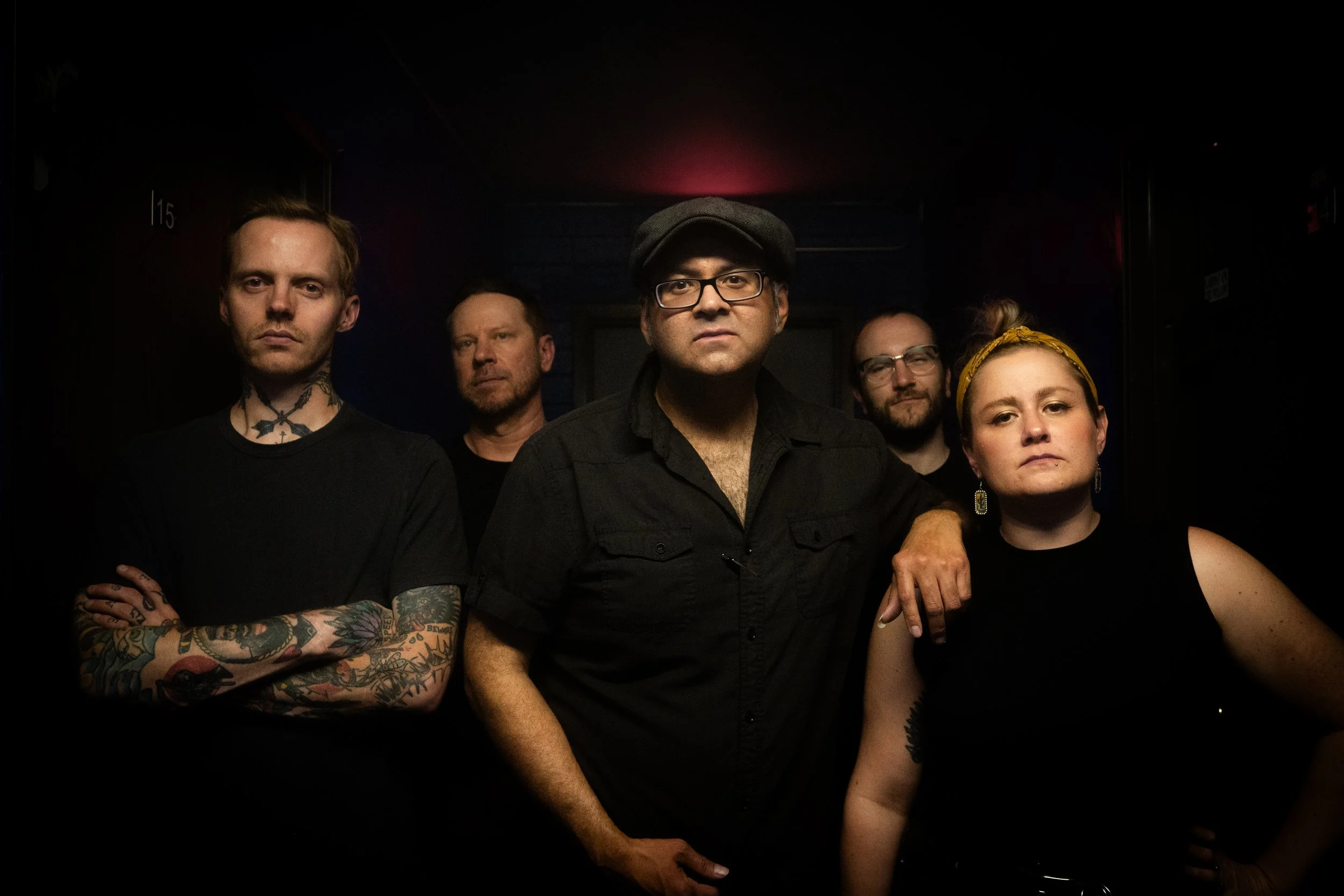 Group of five people posing together in a dark room, with four men and one women, some with tattoos and glasses, wearing black clothing and looking at the camera.