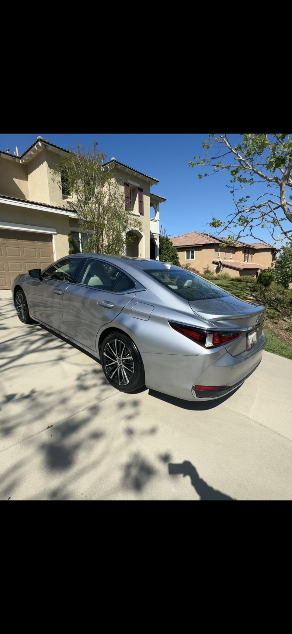 A silver luxury sedan car parked on a driveway in front of a beige house with a brown garage door. The house has trees and shrubs in the yard, and clear blue sky overhead.