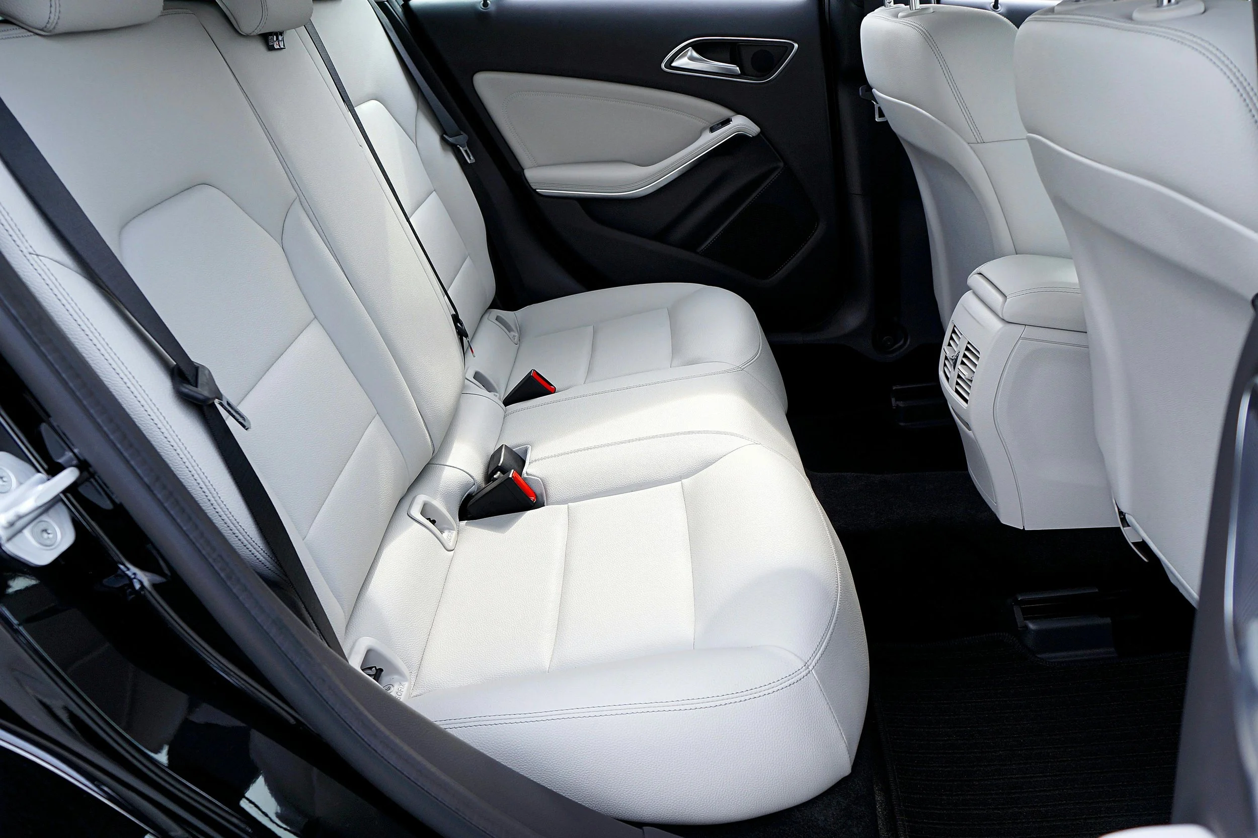 The image shows the rear interior of a car with white leather seats, black seatbelt straps, and a center air conditioning vent at the back of the front seats.