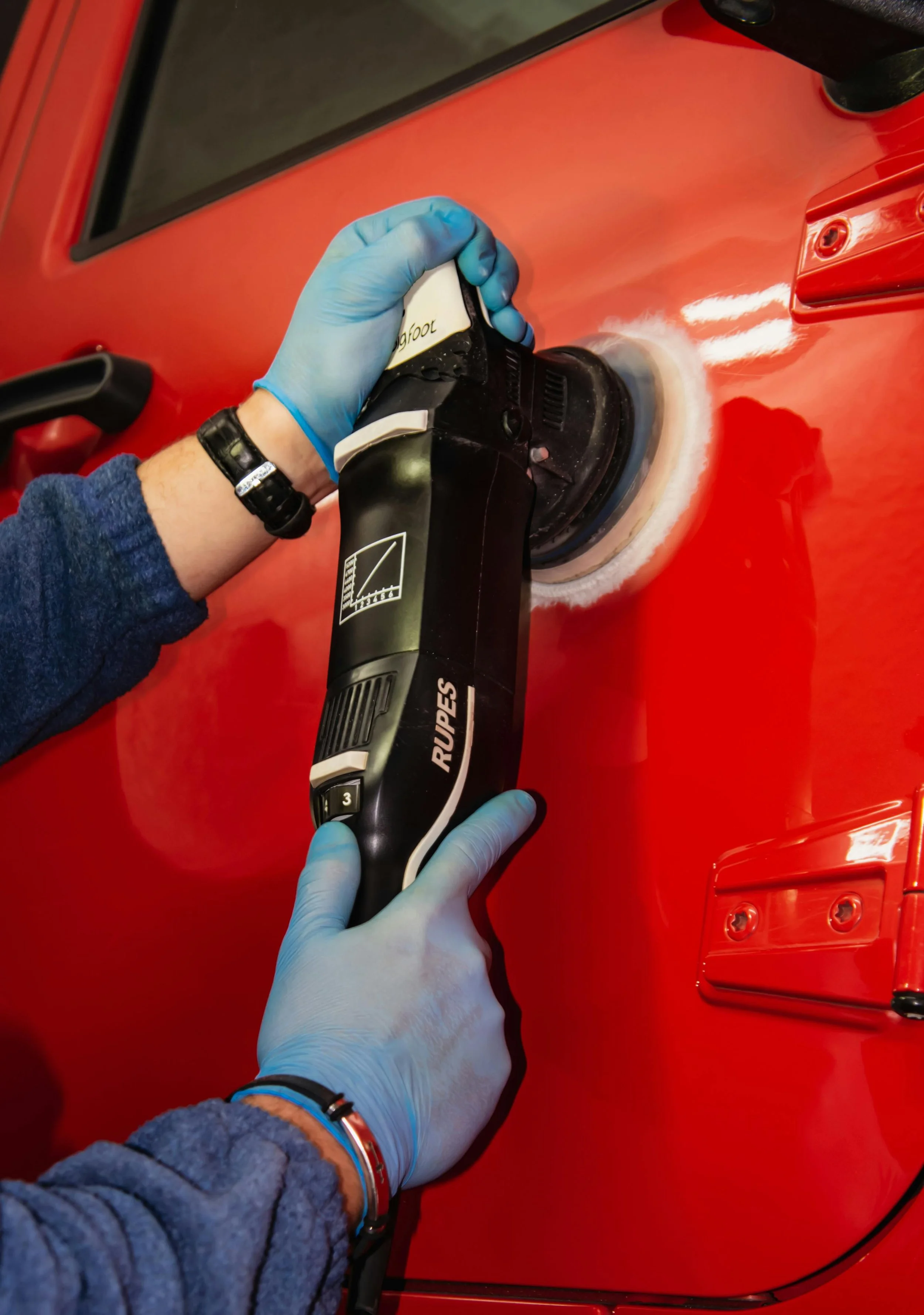 A person wearing a blue glove handling a black and white robot with the brand 'RUPES' on it, polishing a red surface with a polishing machine.