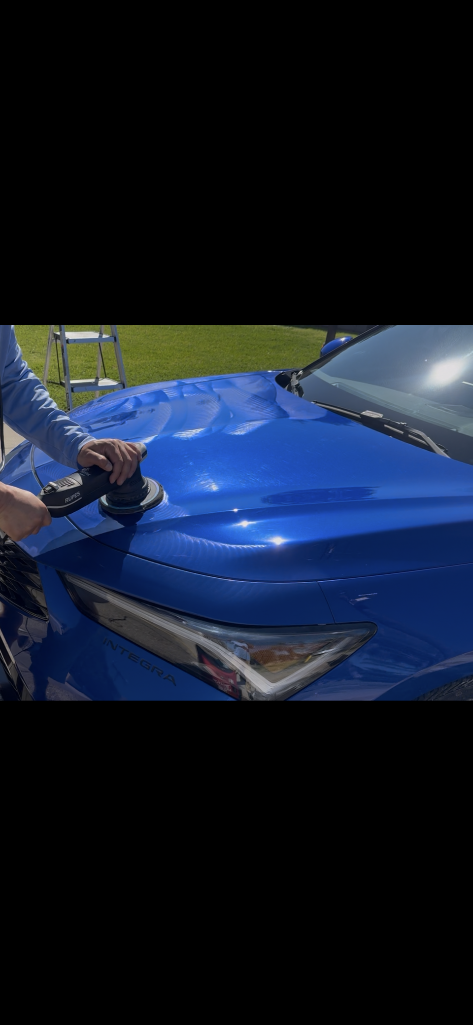 Person using a handheld electric buffer or polisher on the hood of a blue car, which is parked outdoors on a grassy area