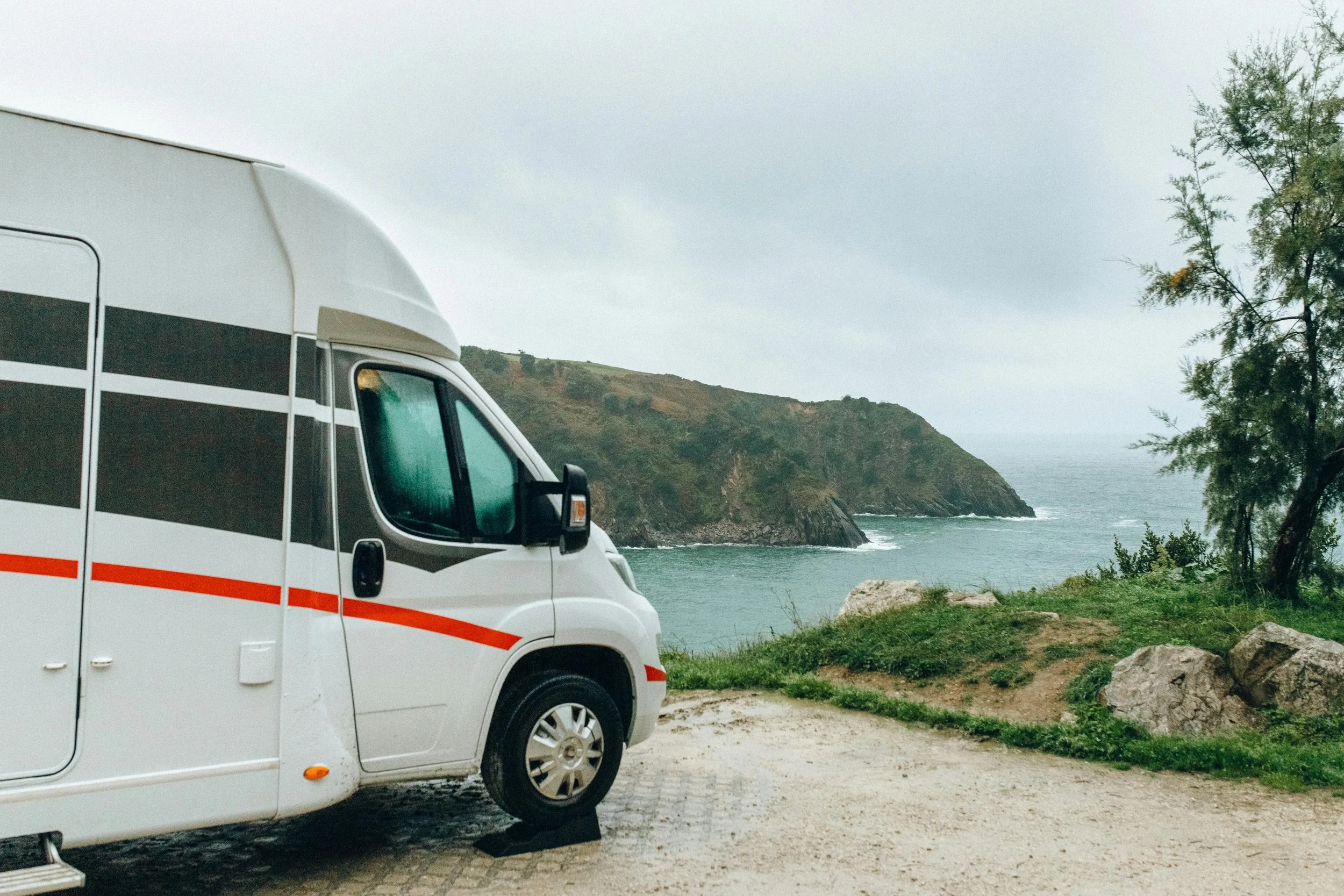 A white camper van parked near the coastline with cliffs and water in the background, partly obscured by trees.