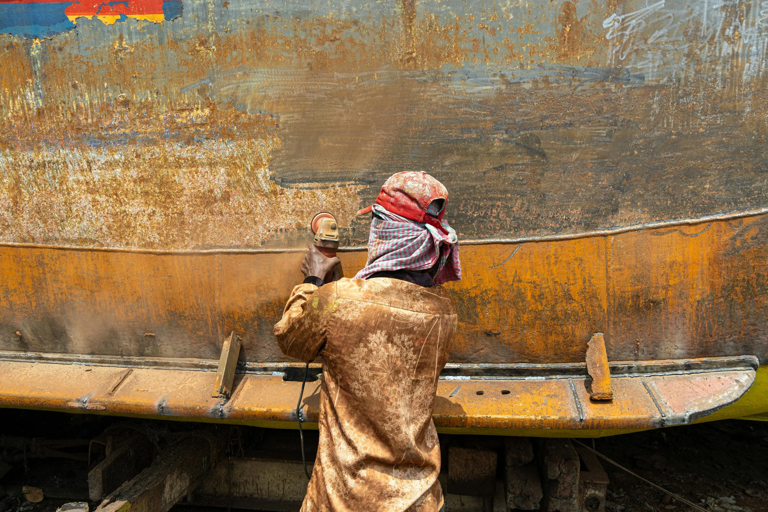 A worker wearing a head covering and long clothing spray-paints a rusty, weathered boat with orange and dark patches. Removal for oxidation