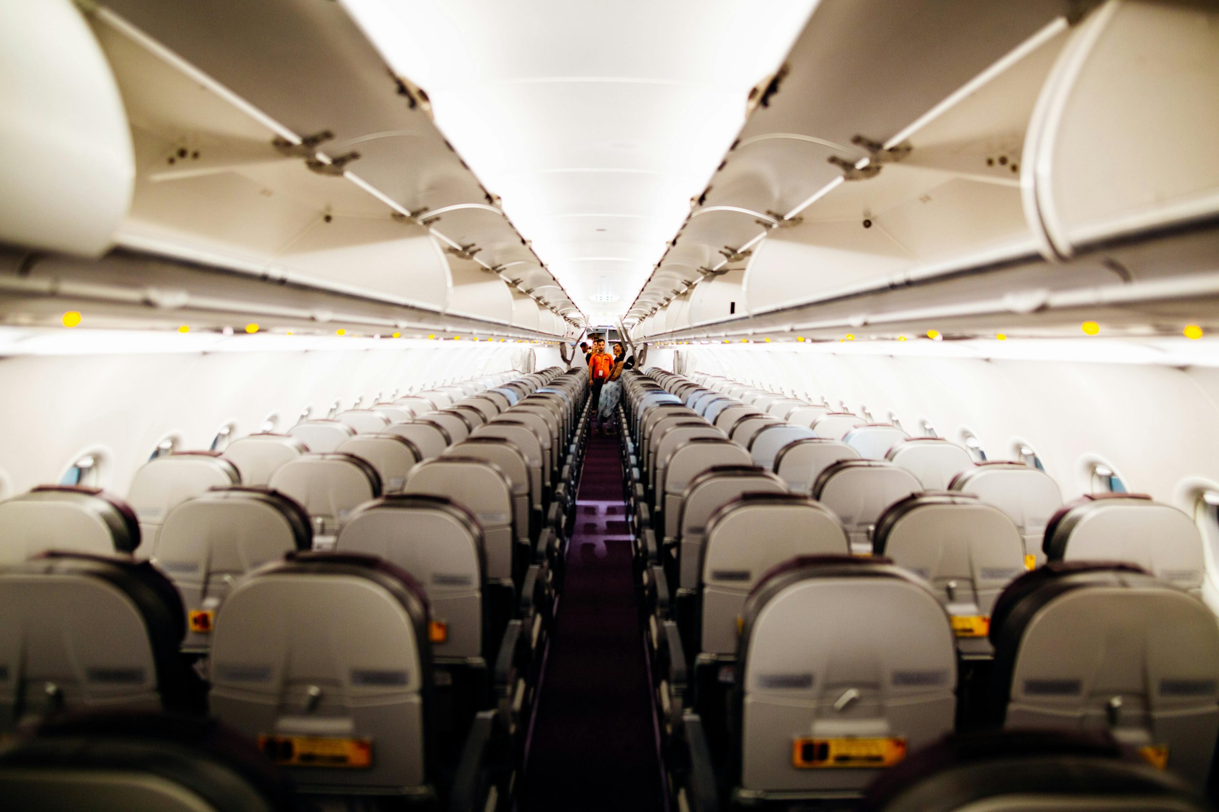 Inside an empty airplane cabin with rows of seats and overhead compartments, a few passengers are standing near the back.