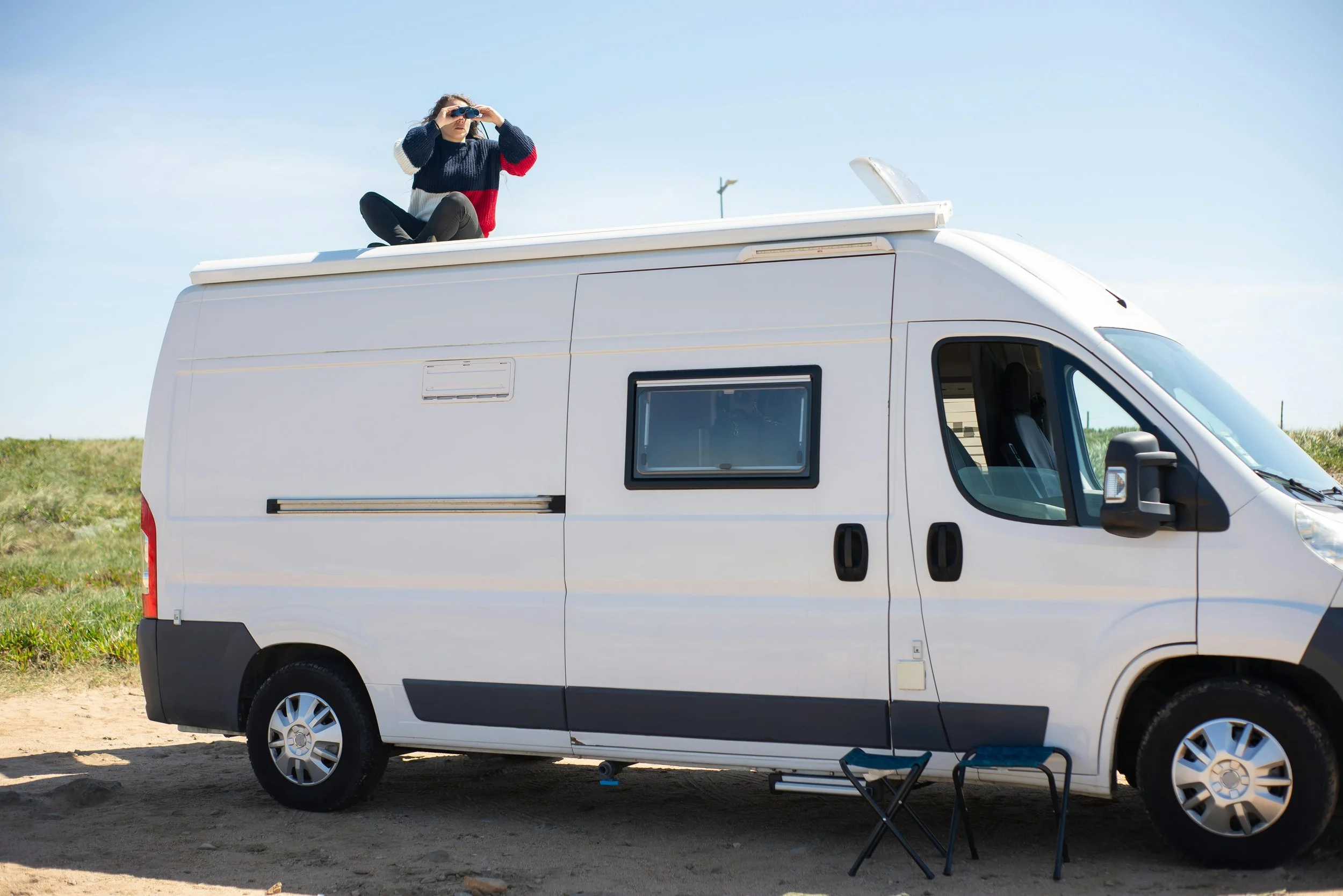 A woman sitting on top of a white camper van in a grassy outdoor area, looking through binoculars on a clear day. Roof Cleaning rv
