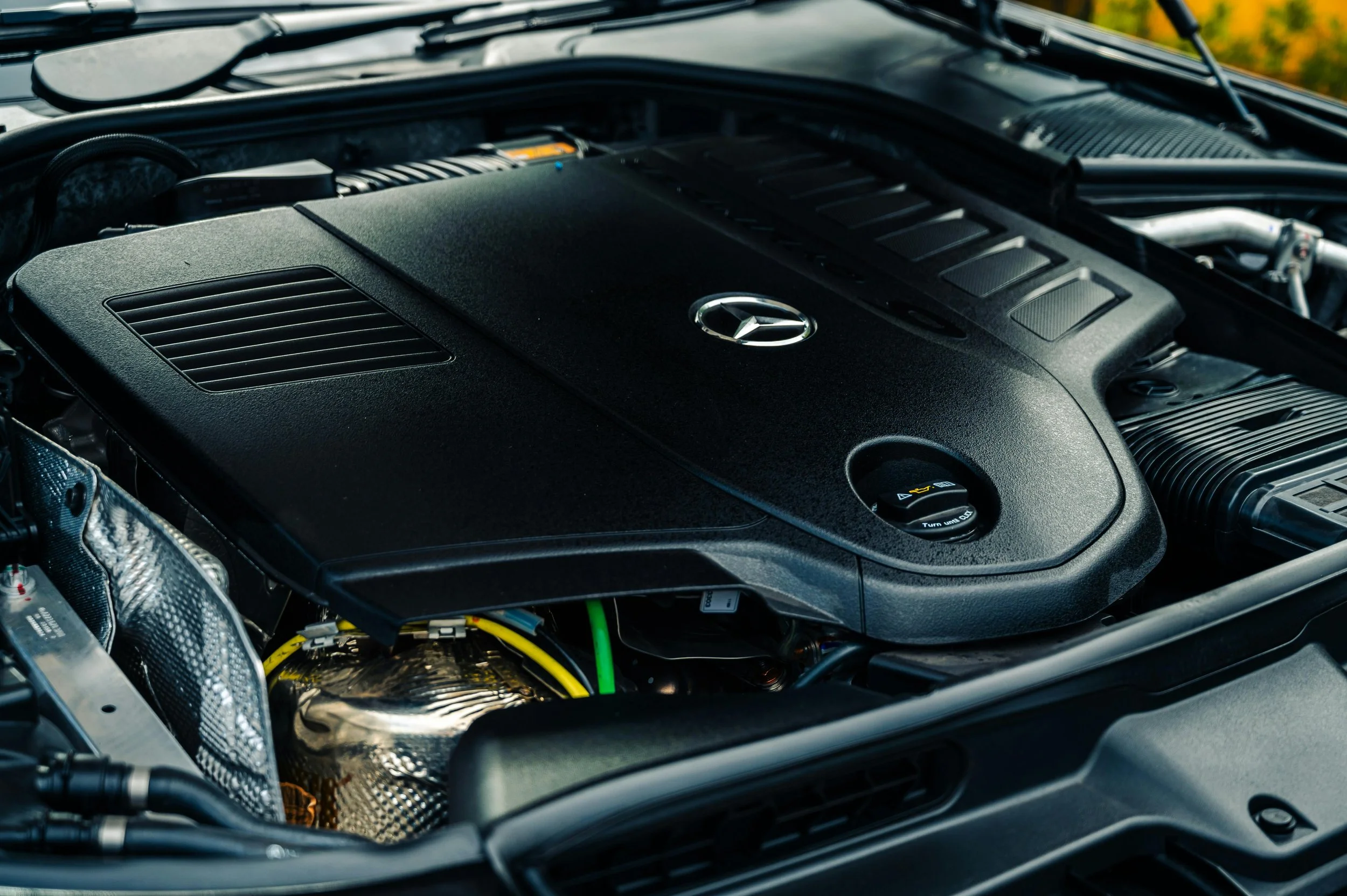 Close-up of a car engine compartment with a Mercedes-Benz logo on the engine cover, featuring black and silver components and some yellow and green wires visible.