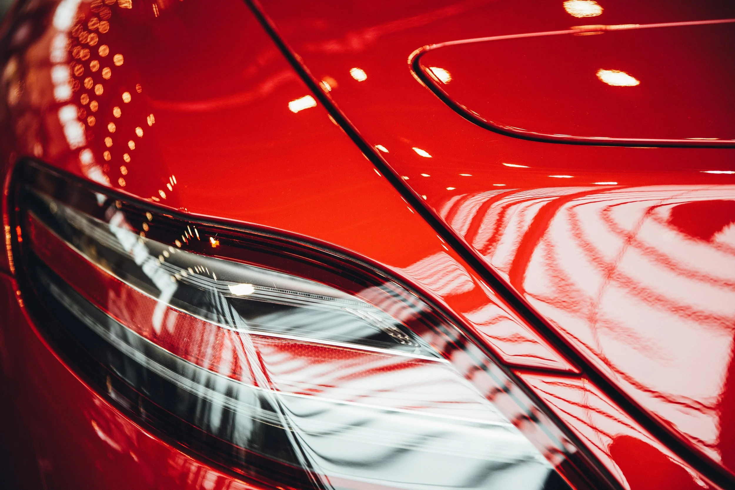 Close-up of a red car's hood and headlight, with reflections of lights on the shiny surface.