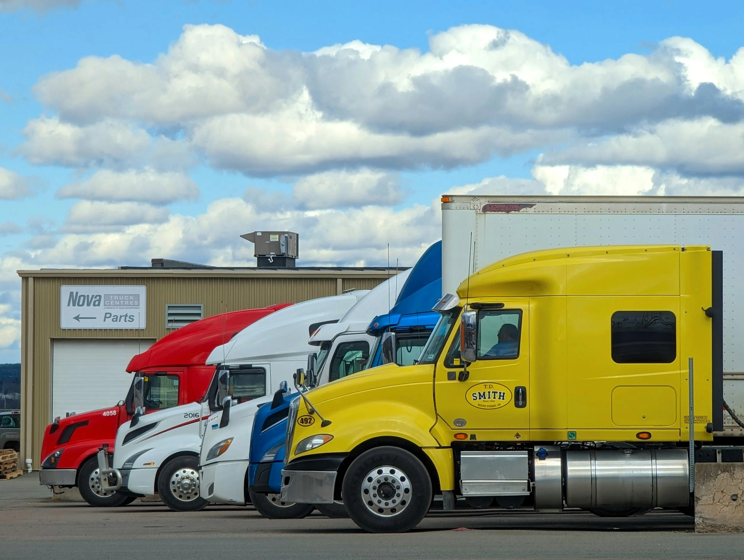 Lineup of semi trucks with yellow, white, red, and blue cabs parked in front of a commercial building under a partly cloudy sky. For Fleet Cleaning.