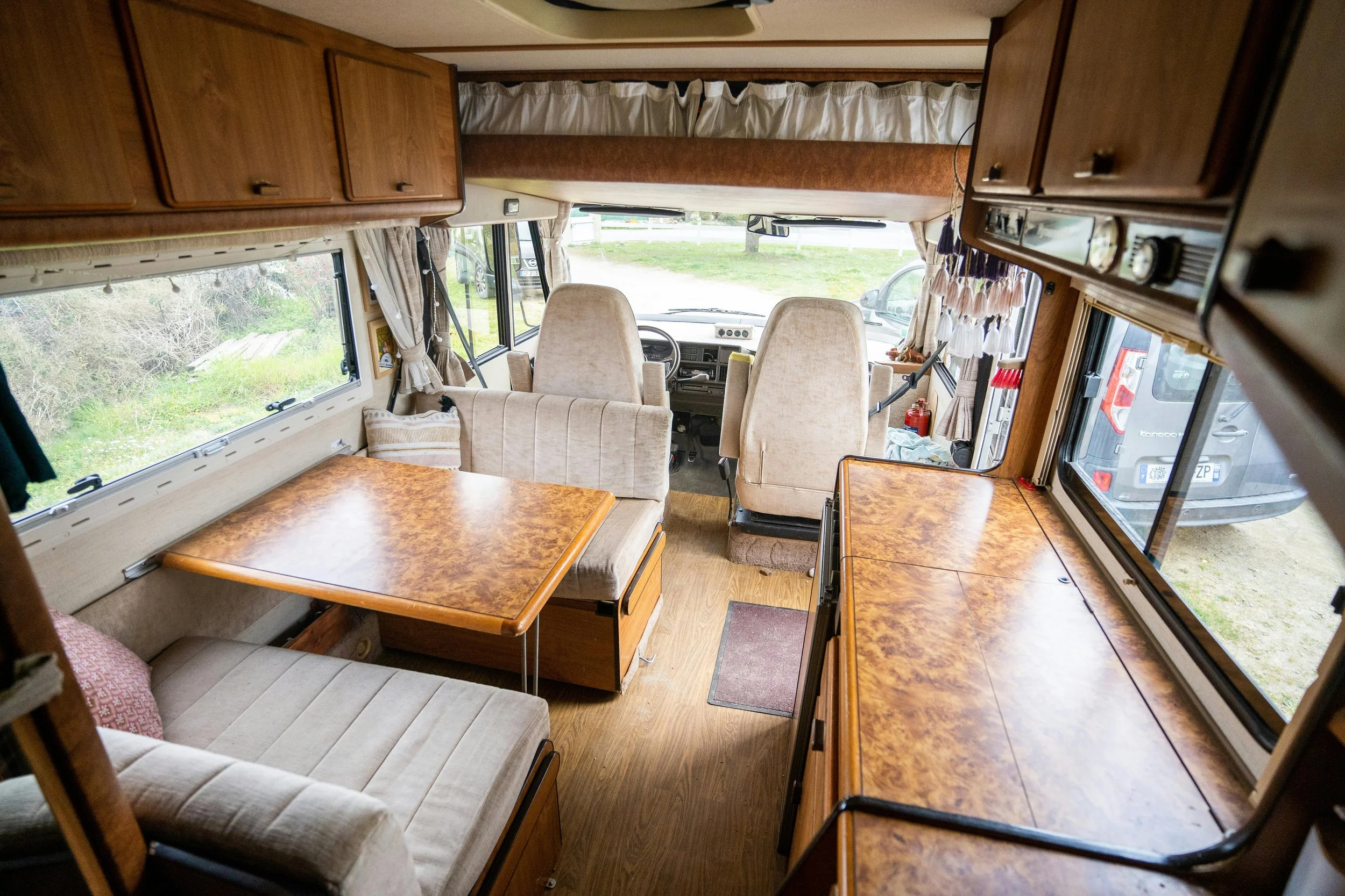 Interior of a vintage camper van showing seating area with beige cushions, wooden tables, large windows with beige curtains, and the driver's cab with two beige seats.