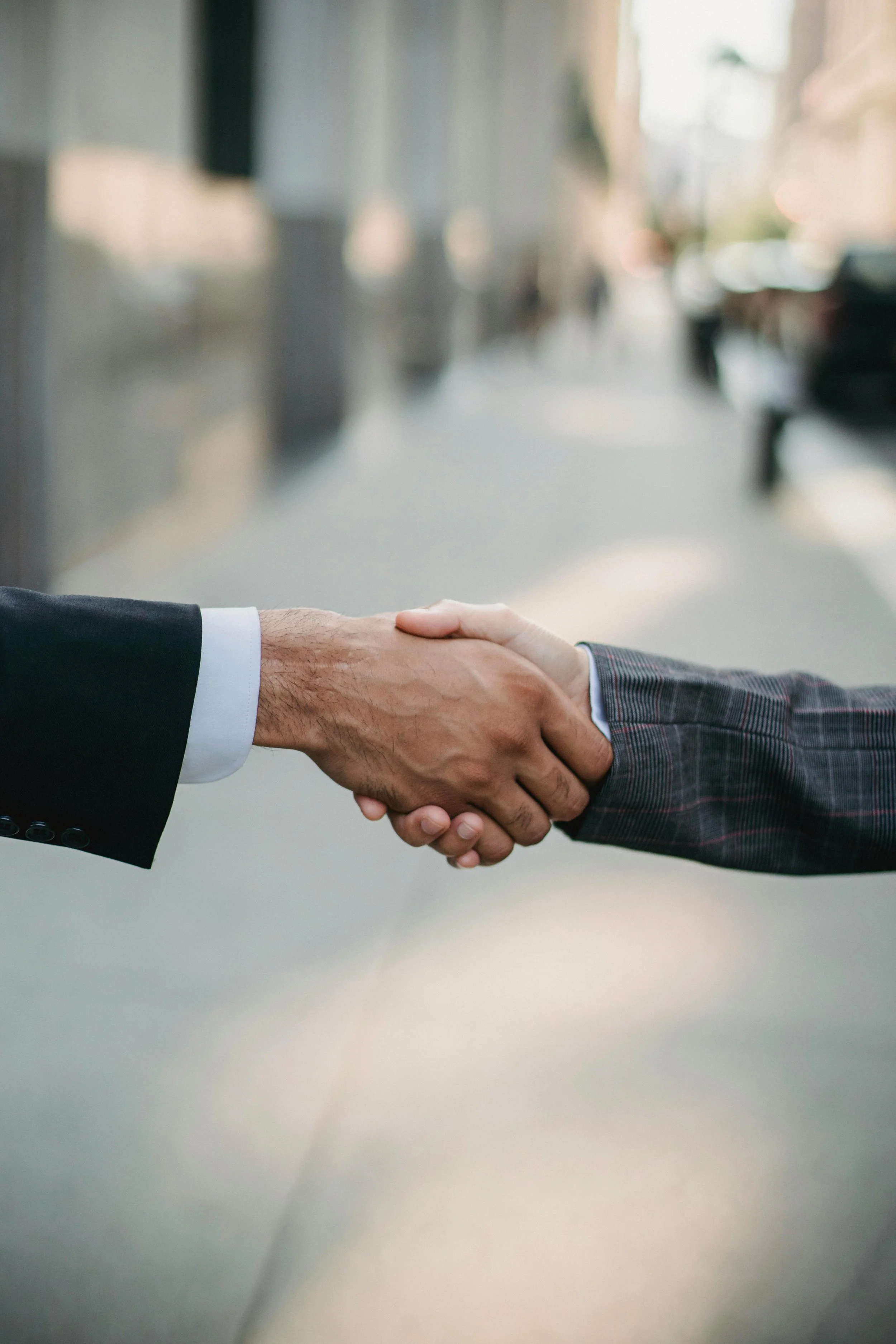 A close-up of two people shaking hands in a professional setting, with blurred outdoor city street background.