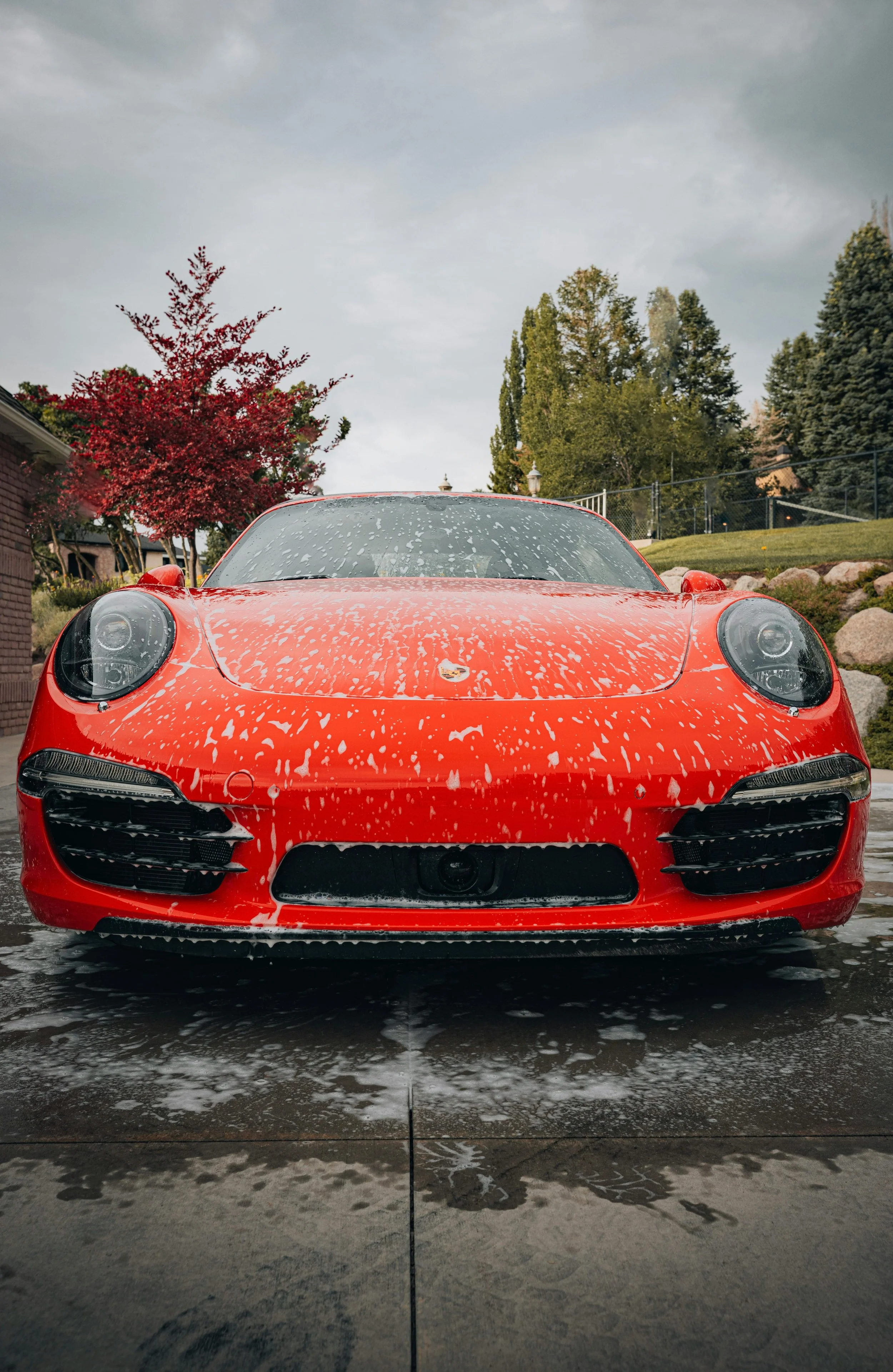 Red sports car covered in soap suds during a car wash, with a wet concrete surface reflecting the vehicle, trees, and cloudy sky in the background.