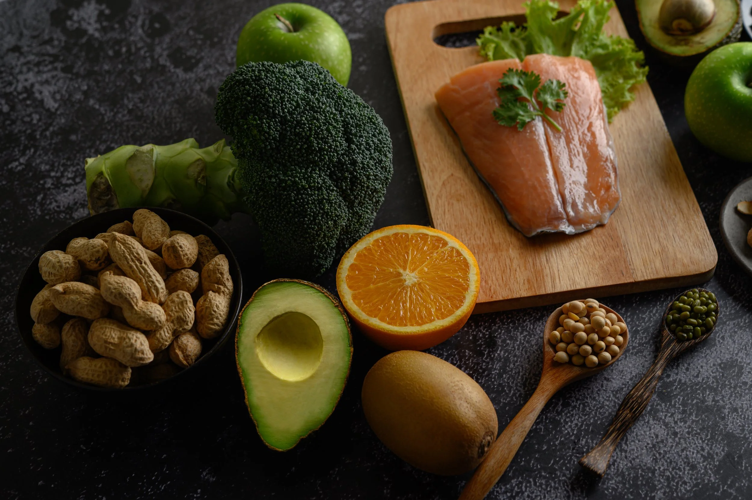 Fresh produce on a black countertop with a wooden cutting board, including broccoli, green apples, an orange, an avocado, and a piece of salmon garnished with cilantro.
