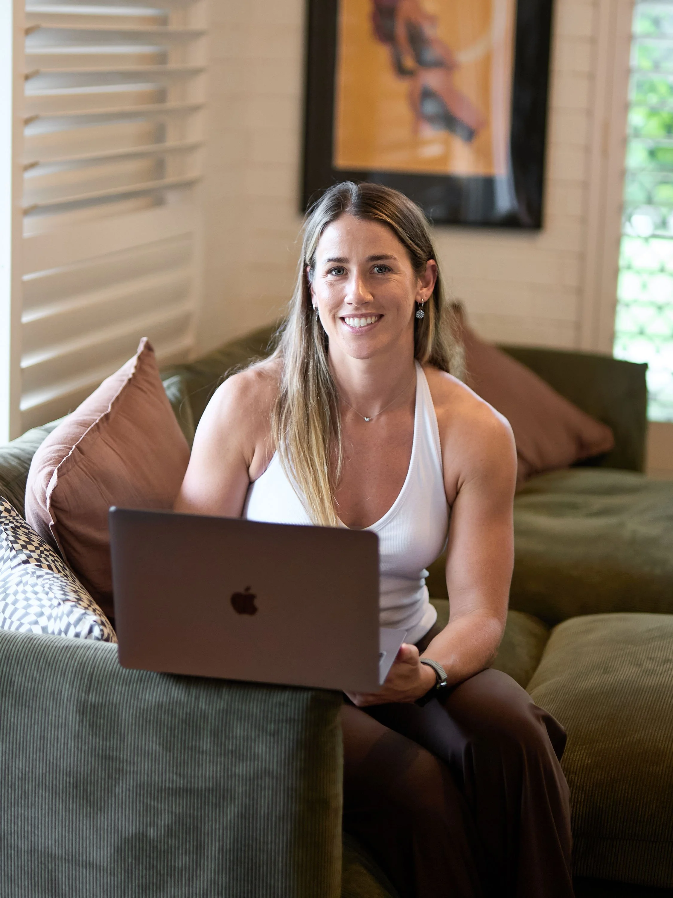 A woman with long hair sitting on a green couch using a laptop, with pillows behind her, in a room with wooden walls and a framed artwork