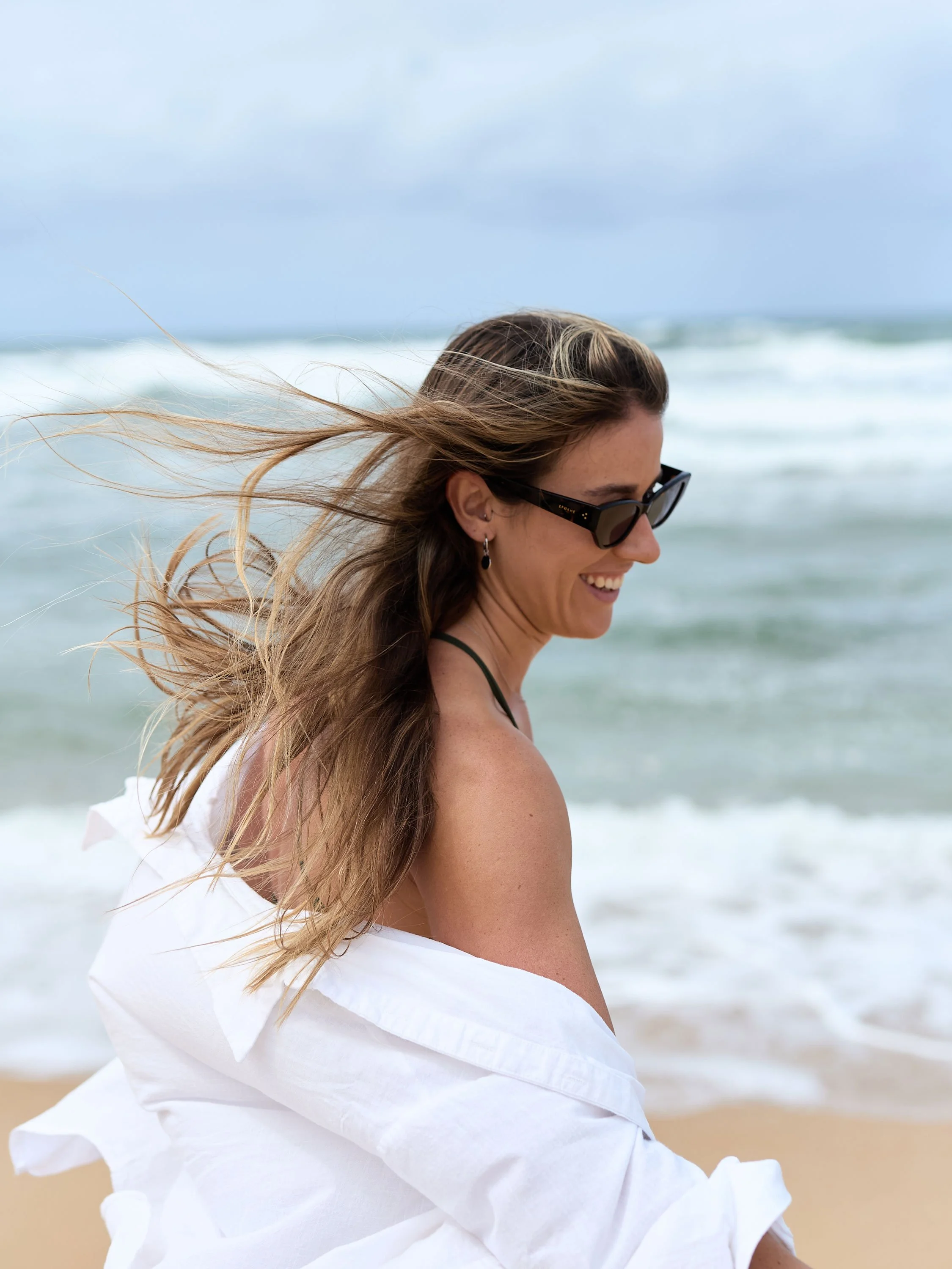 Woman with long hair wearing sunglasses and a white dress, smiling on a beach with ocean waves in the background.