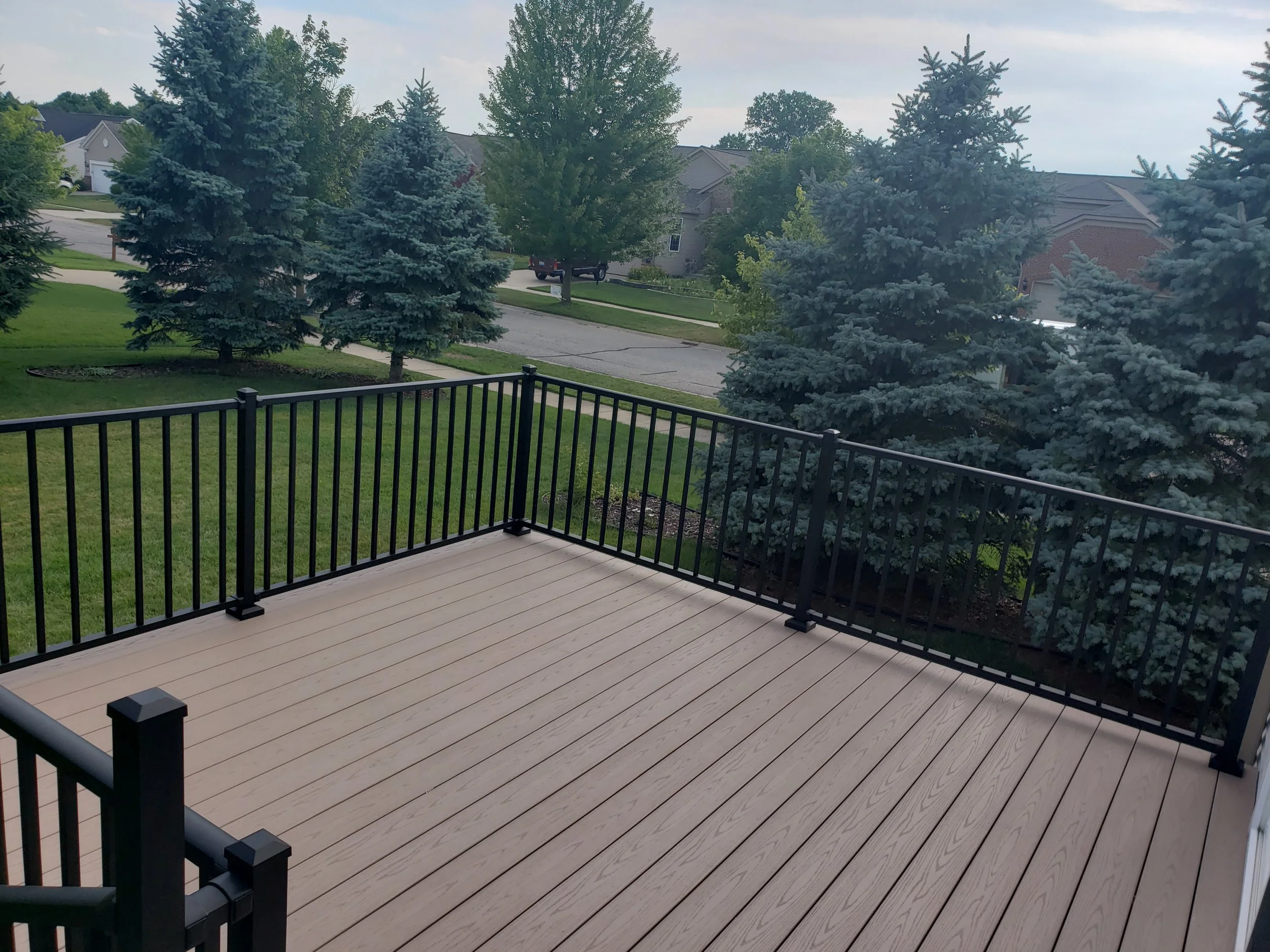 A backyard view from a wooden deck with black railings, overlooking a neighborhood street lined with trees and houses, under a cloudy sky.