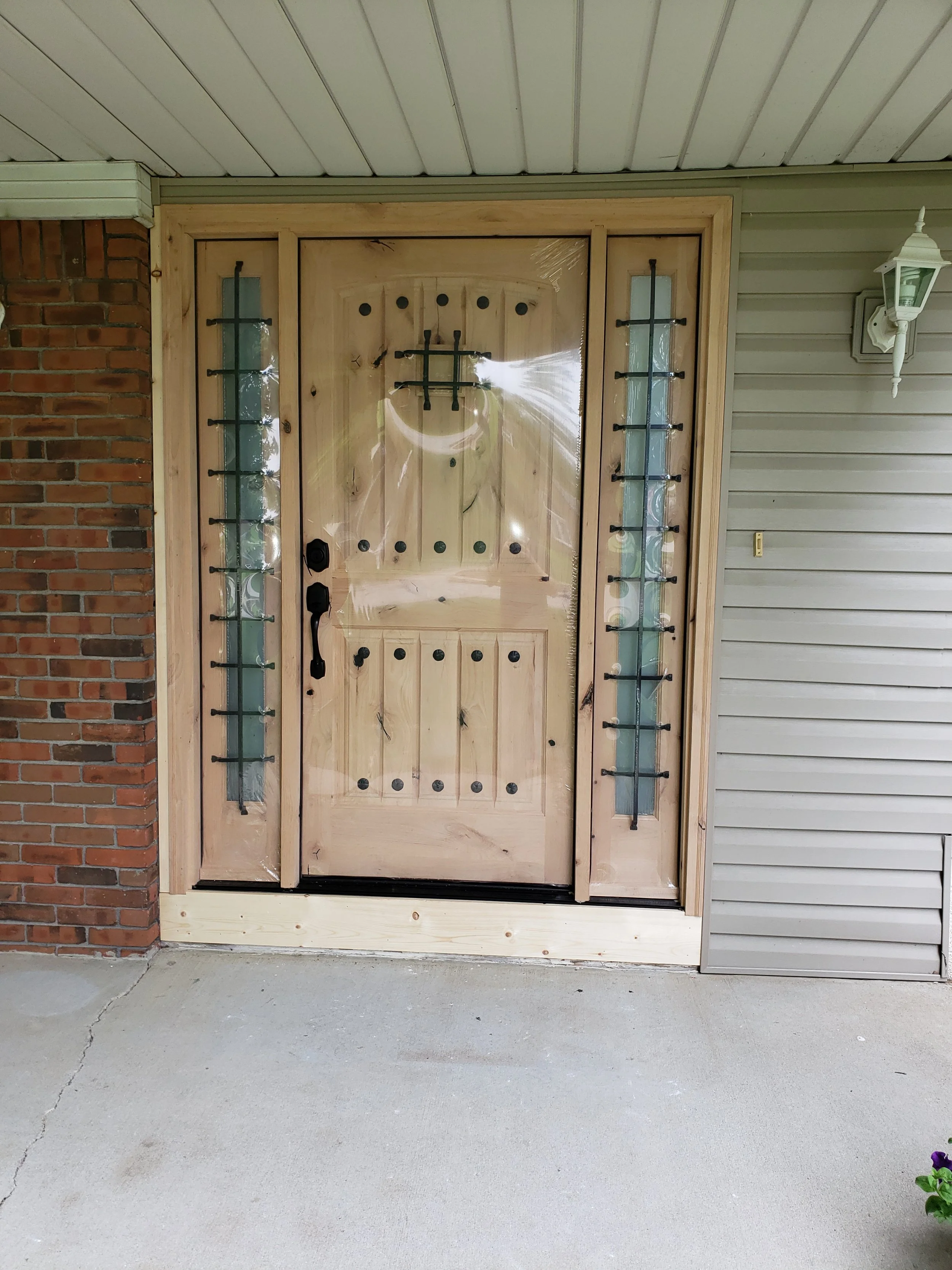Front view of a house entrance with a new wooden door and sidelights, surrounded by brick and siding, with a porch light on the right side and a concrete porch in the foreground.