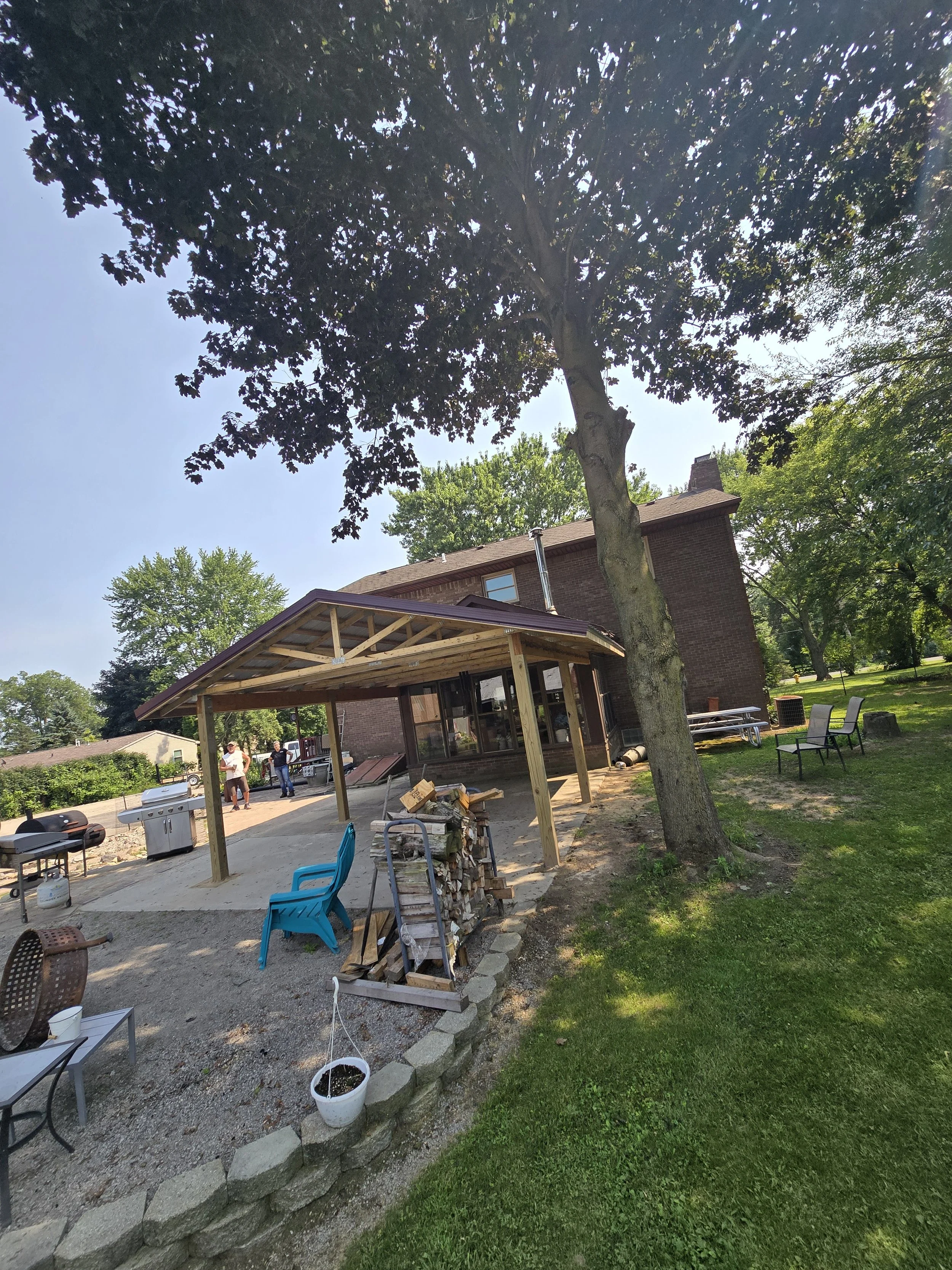 Backyard with a large tree, a brick house, people, outdoor furniture, and a construction area with wood and pavers.