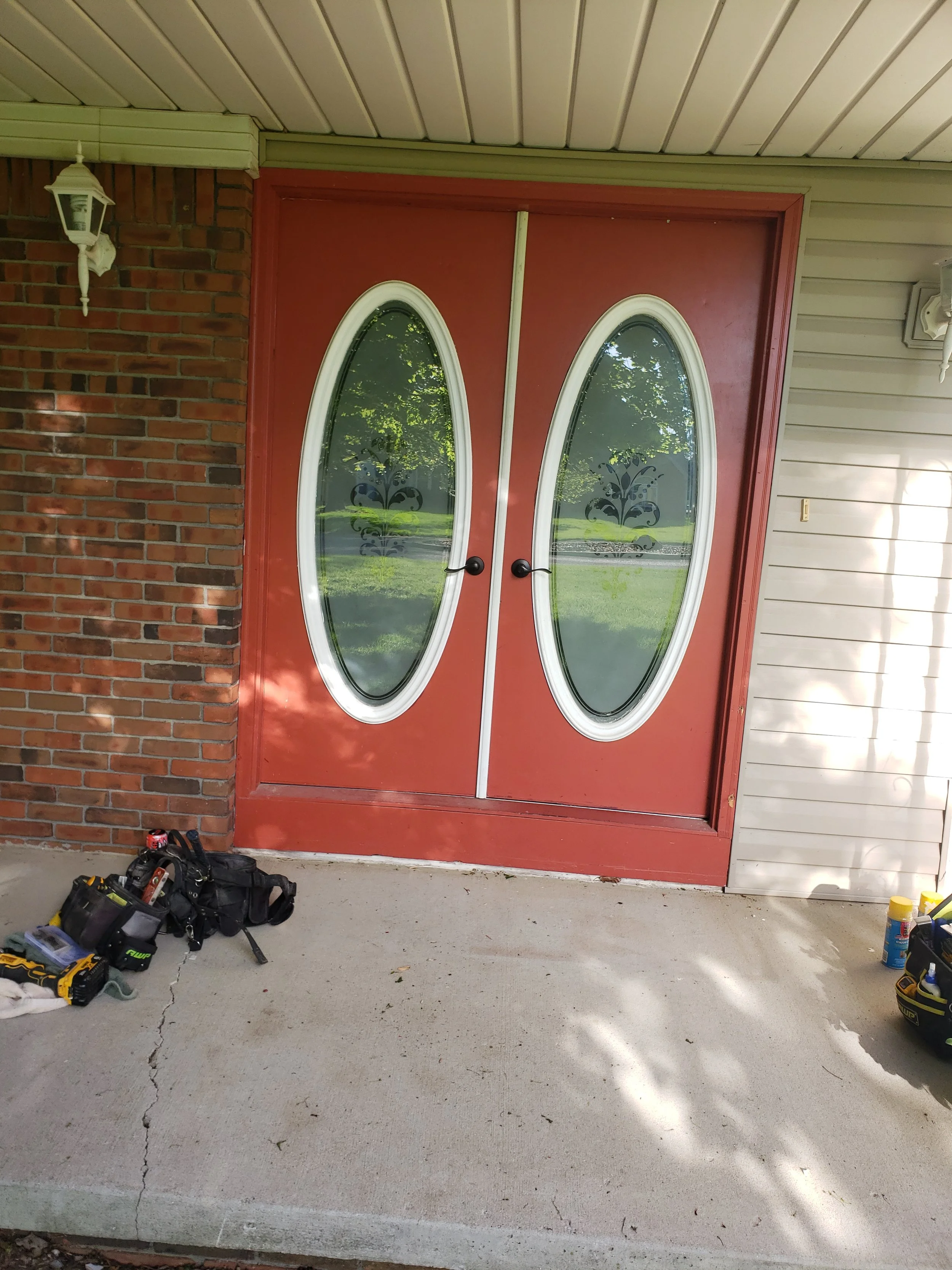 Front porch with red double doors with oval glass panels, brick wall on the left, siding on the right, and outdoor lighting fixtures. Tools and a backpack are on the concrete porch.