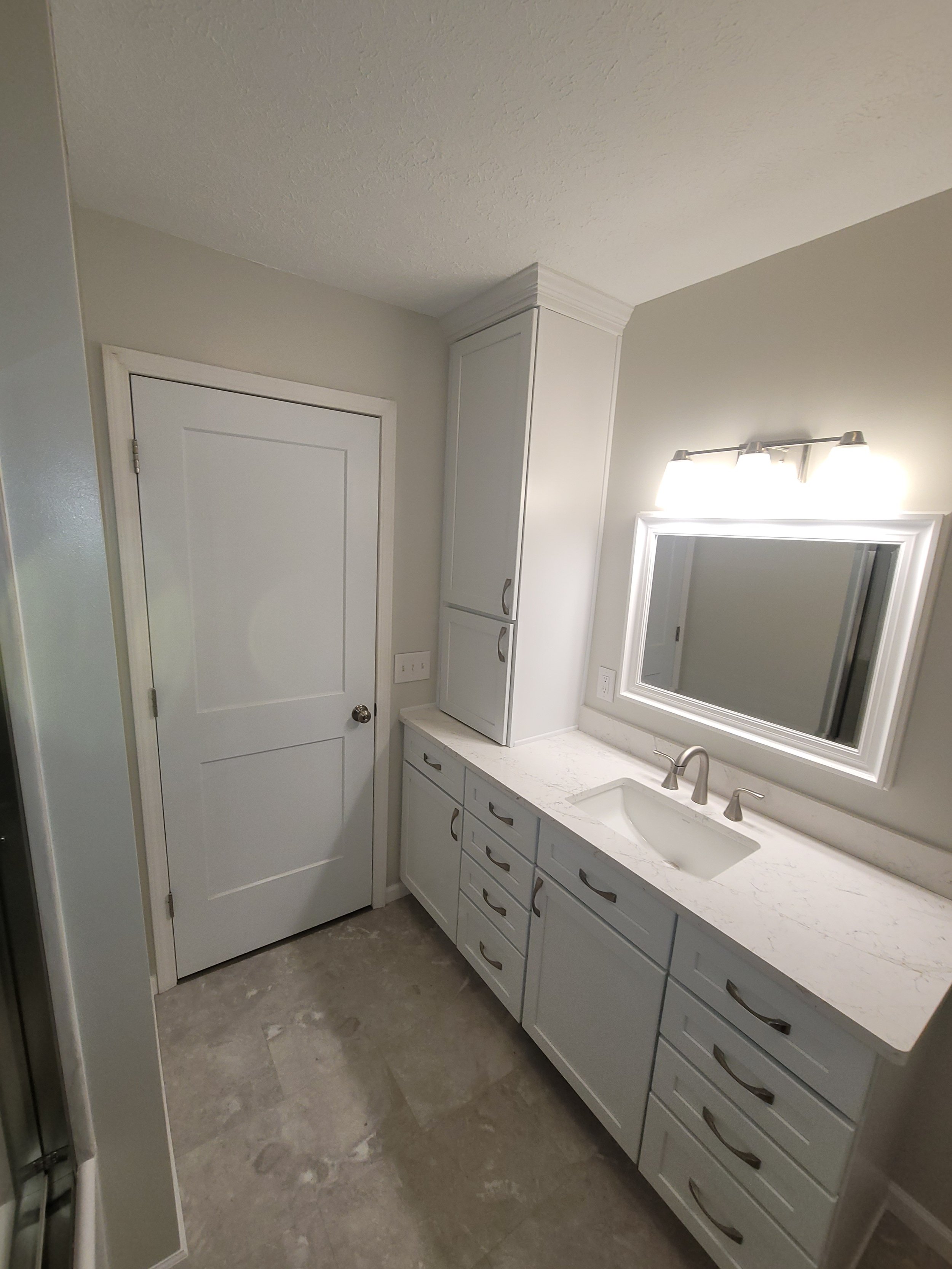 A bathroom vanity area with a marble countertop, a single sink, and a mirror above it. There is a light fixture with three bulbs above the mirror. To the left, there is a closed white door and a tall white cabinet with drawers and handles below. The floor is tiled in a grayish color.