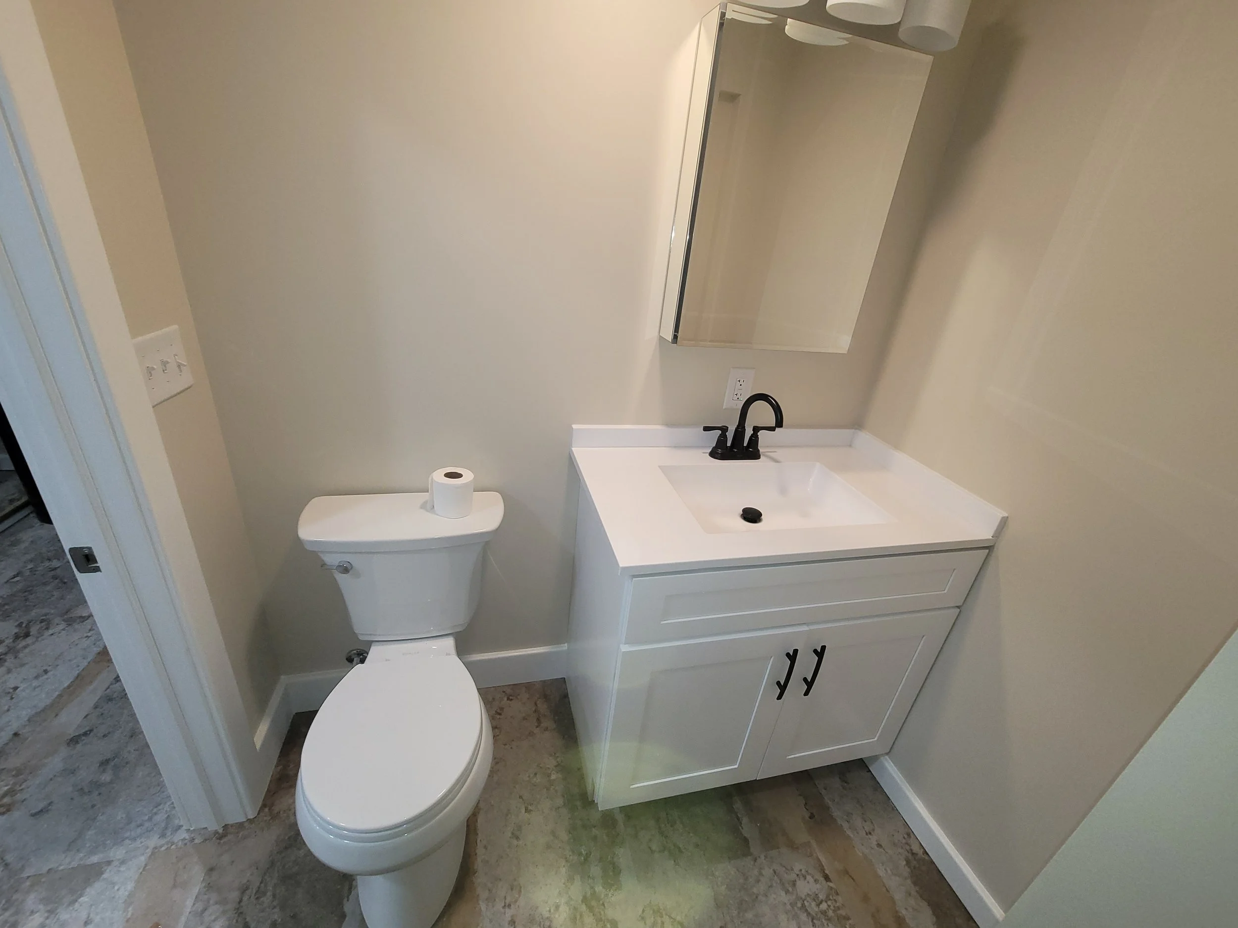 A small bathroom with light gray walls, a white toilet with a roll of toilet paper on the tank, a white sink with a black faucet, a mirror, and a white cabinet with black hooks at the bottom.
